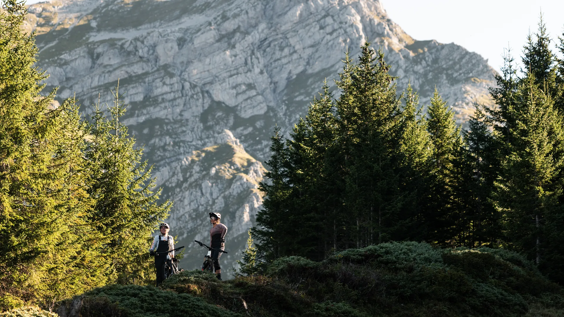 Zwei Bikerinnen mit ihren Bikes stehen auf einer Lichtung, umgeben von Tannen, imposantes Felsmassiv im Hintergrund