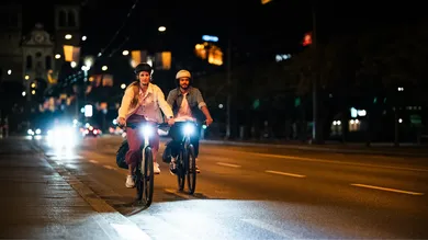 Zwei Fahrer:innen mit beleuchtetem E-Bike bei Nacht in der Stadt, Frontalansicht