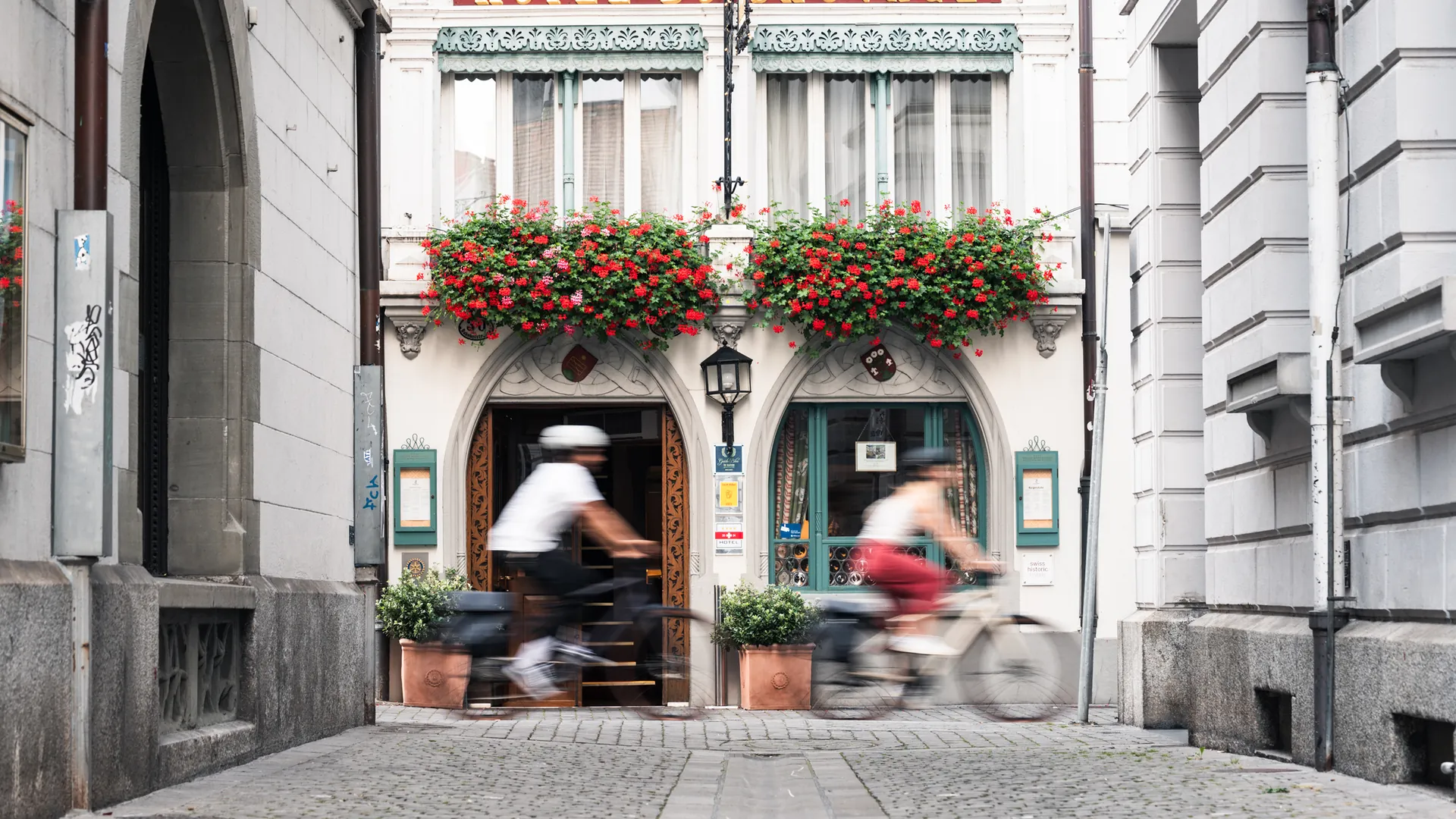 Zwei Fahrer:innen in Bewegungsunschärfe vor historischer Altstadtfassade mit Blumenschmuck
