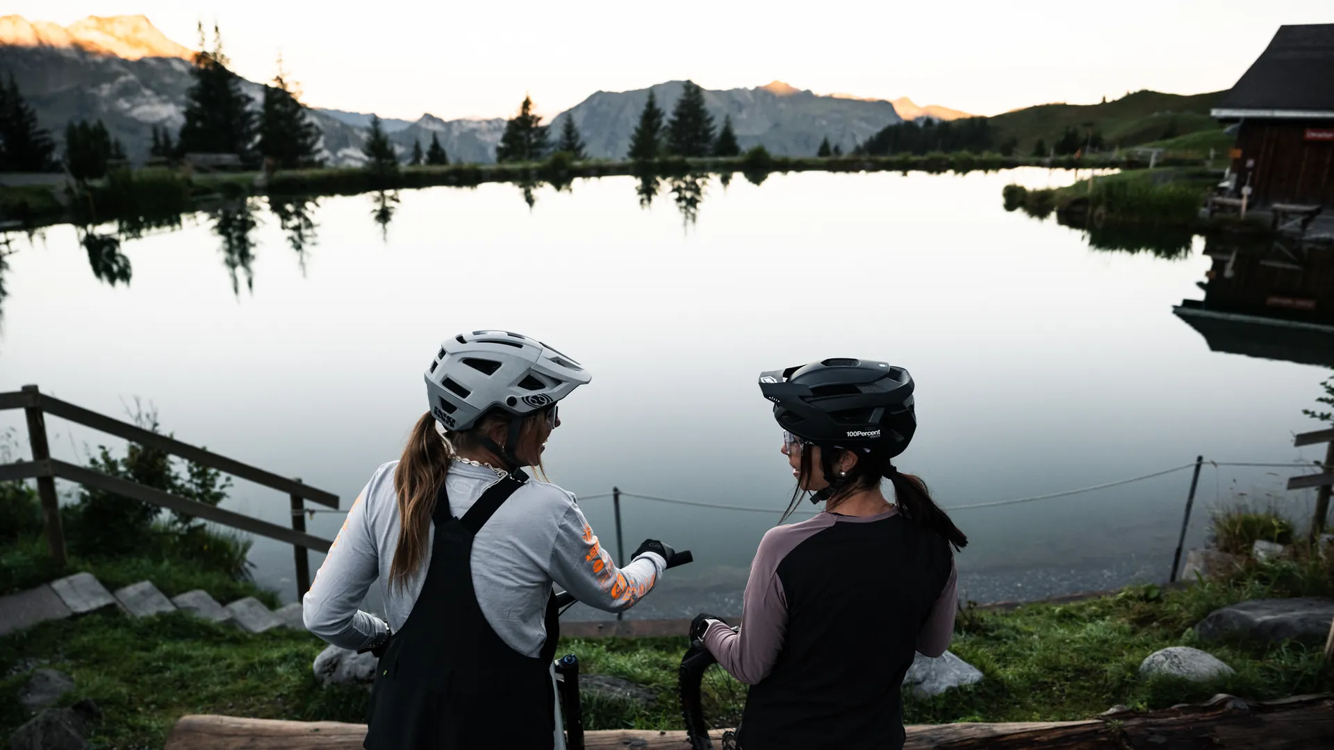 Zwei Bikerinnen mit Helm stehen im Gespräch vor einem spiegelglatten Bergsee bei Abenddämmerung, Alpenpanorama im Hintergrund
