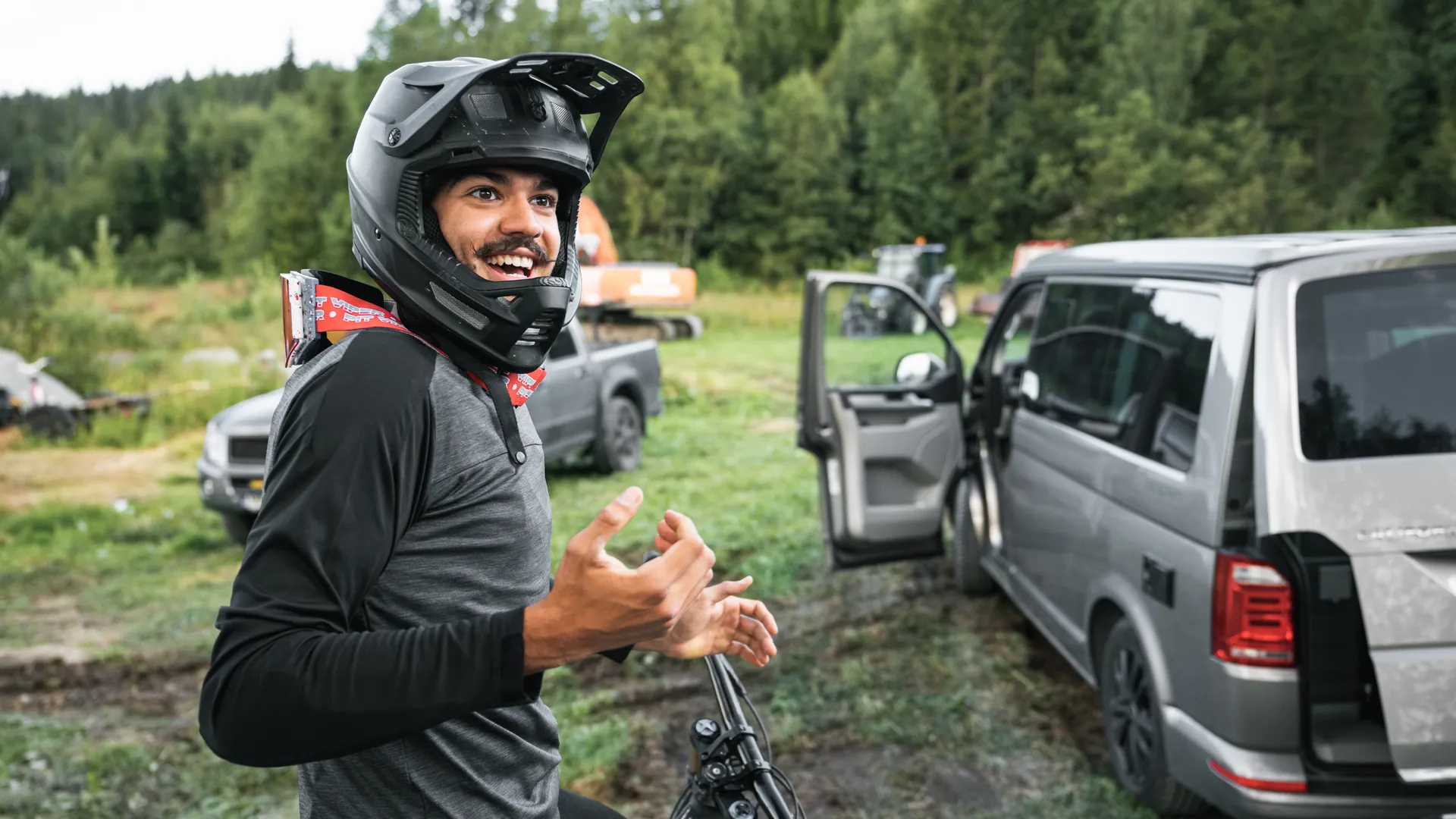 Lachender Mann mit Fullface-Helm gestikuliert vor Fahrzeugen auf einer Wies
