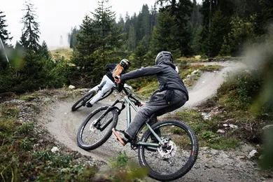 Zwei Fahrer:innen in enger Kurve auf Waldtrail, Vorderrad im Wheelie