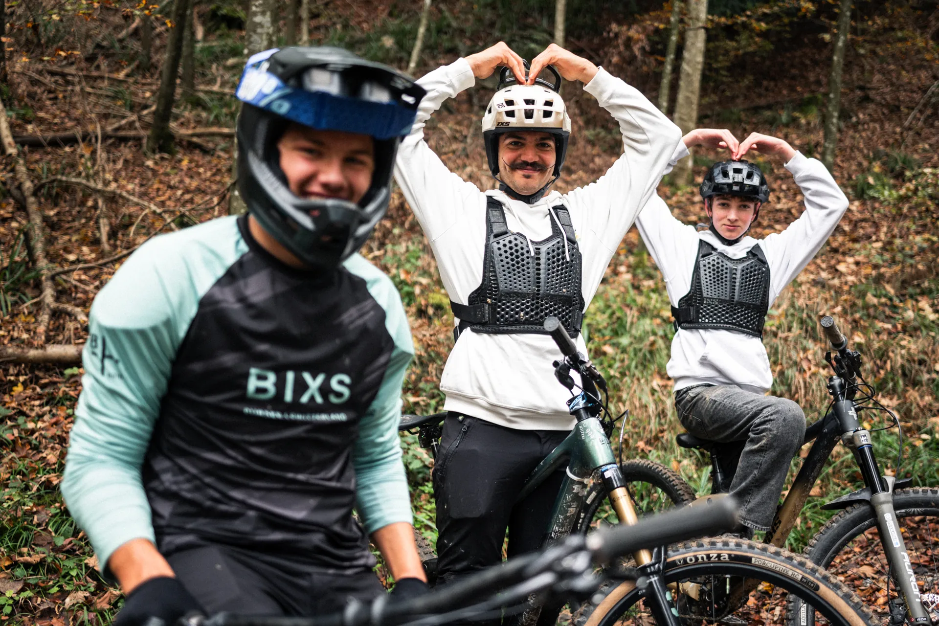Drei BIXS Fahrer mit Fullface-Helmen und Protektoren formen Herzzeichen mit den Händen im herbstlichen Wald
