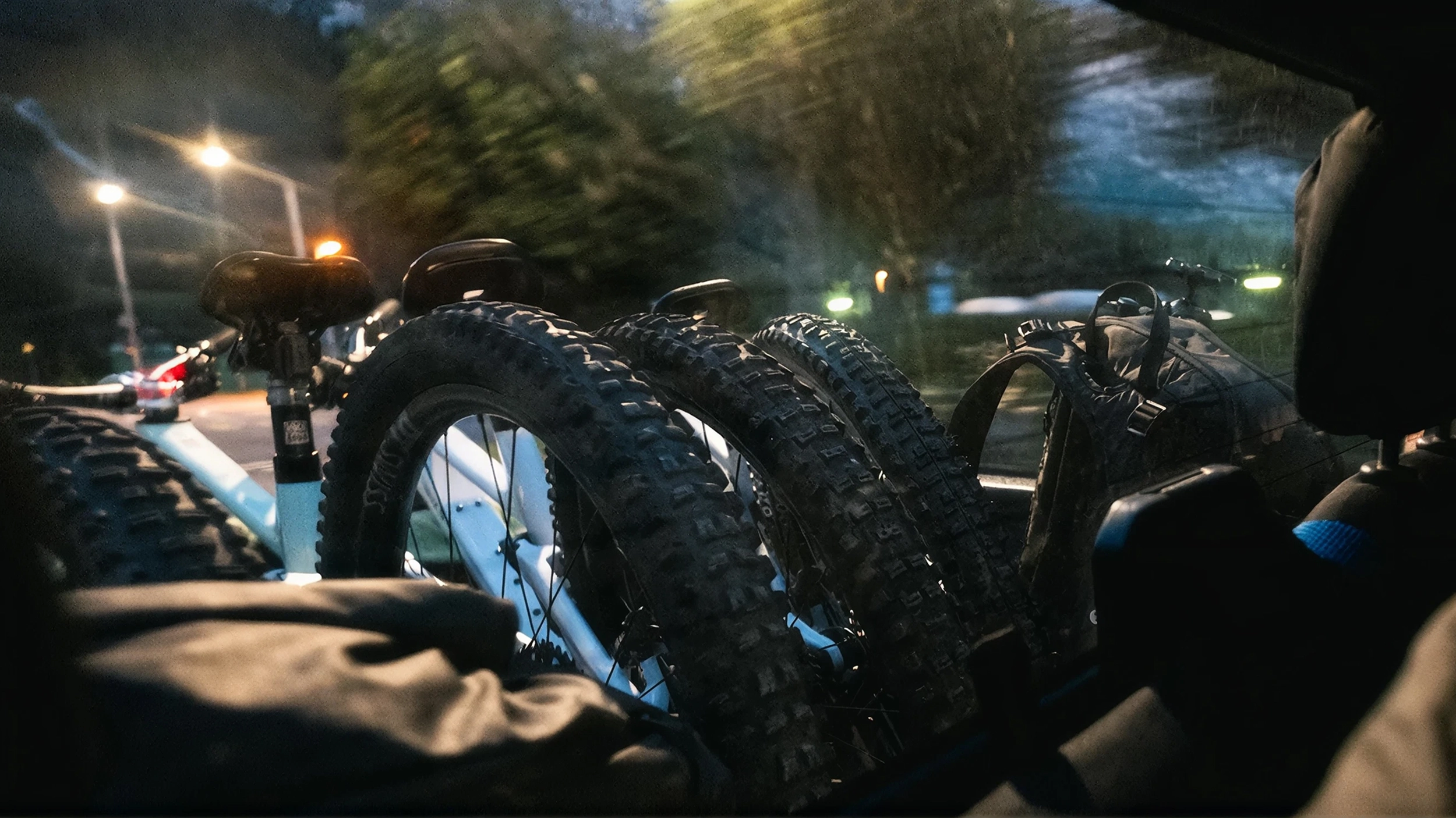 Blick aus dem Kofferraum auf mehrere transportierte Bikes mit groben Stollenreifen, nächtliche Strassenszene mit Lichtern im Hintergrund