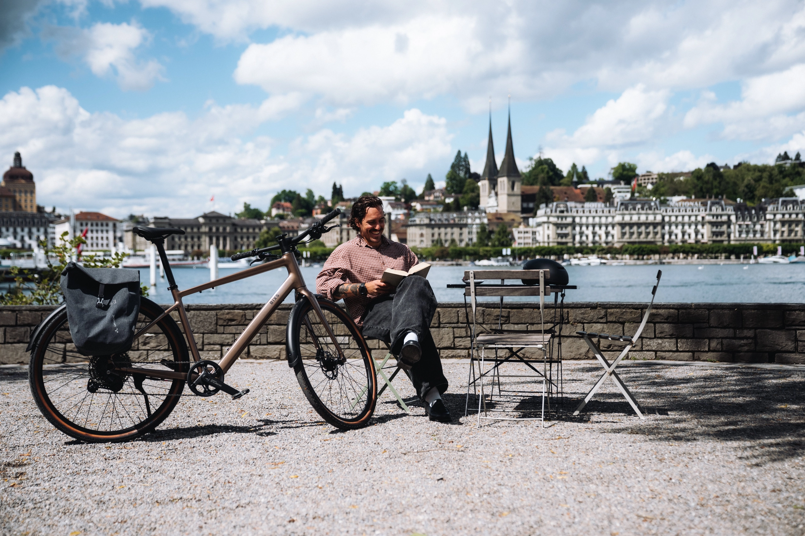 Mann liest lächelnd ein Buch neben seinem District E-Bike am Seeufer, Luzerner Altstadt-Silhouette mit Hofkirche im Hintergrund