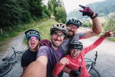 Vier Mountainbikerinnen und Mountainbiker jubeln lachend in die Kamera für ein Gruppenselfie auf einem Kiesweg