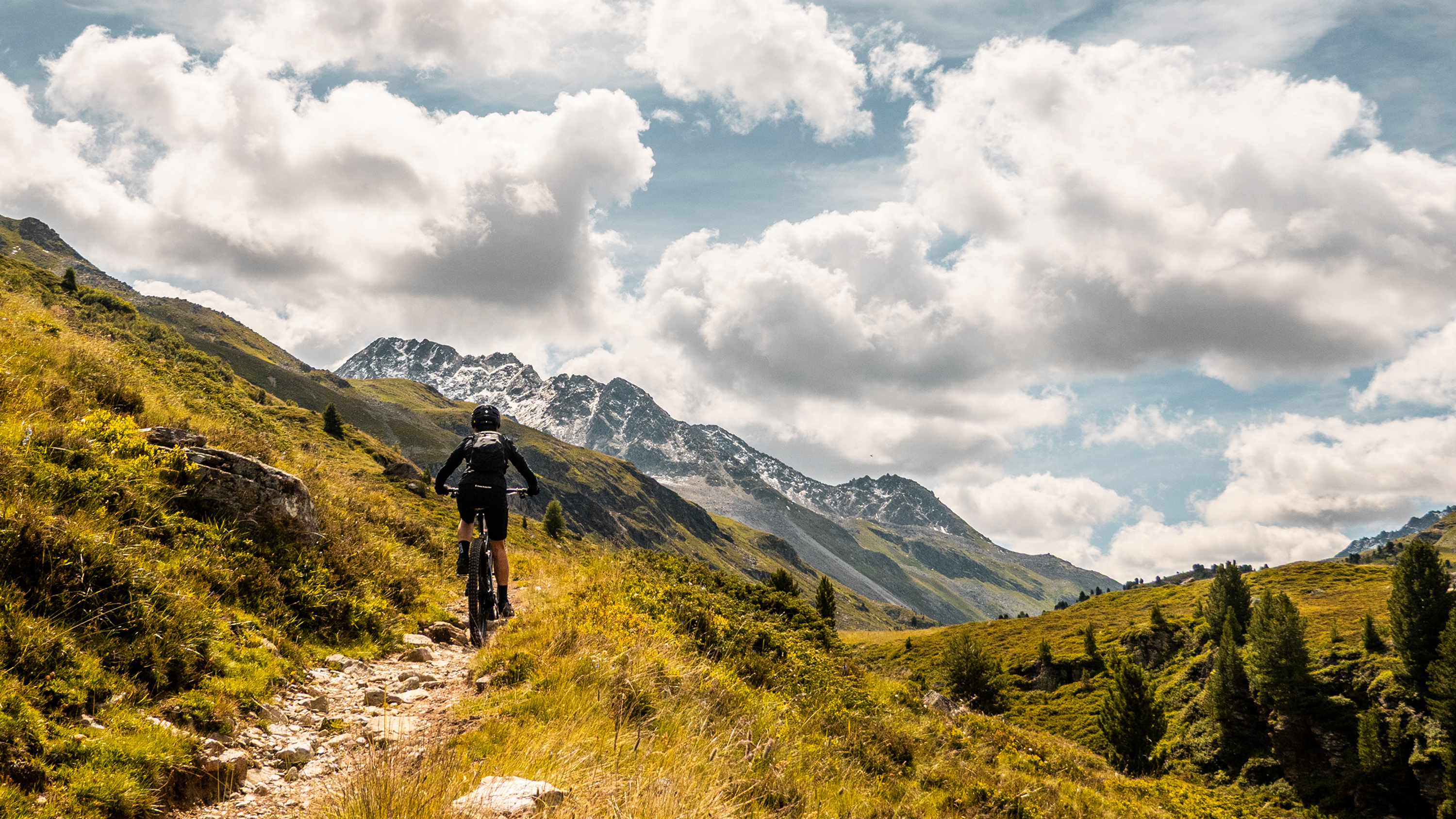 Eine Person, die mit dem Mountainbike auf einem felsigen Weg durch üppig grüne Hügel fährt, im Hintergrund sind schneebedeckte Berge und ein blauer Himmel mit Wolken zu sehen.