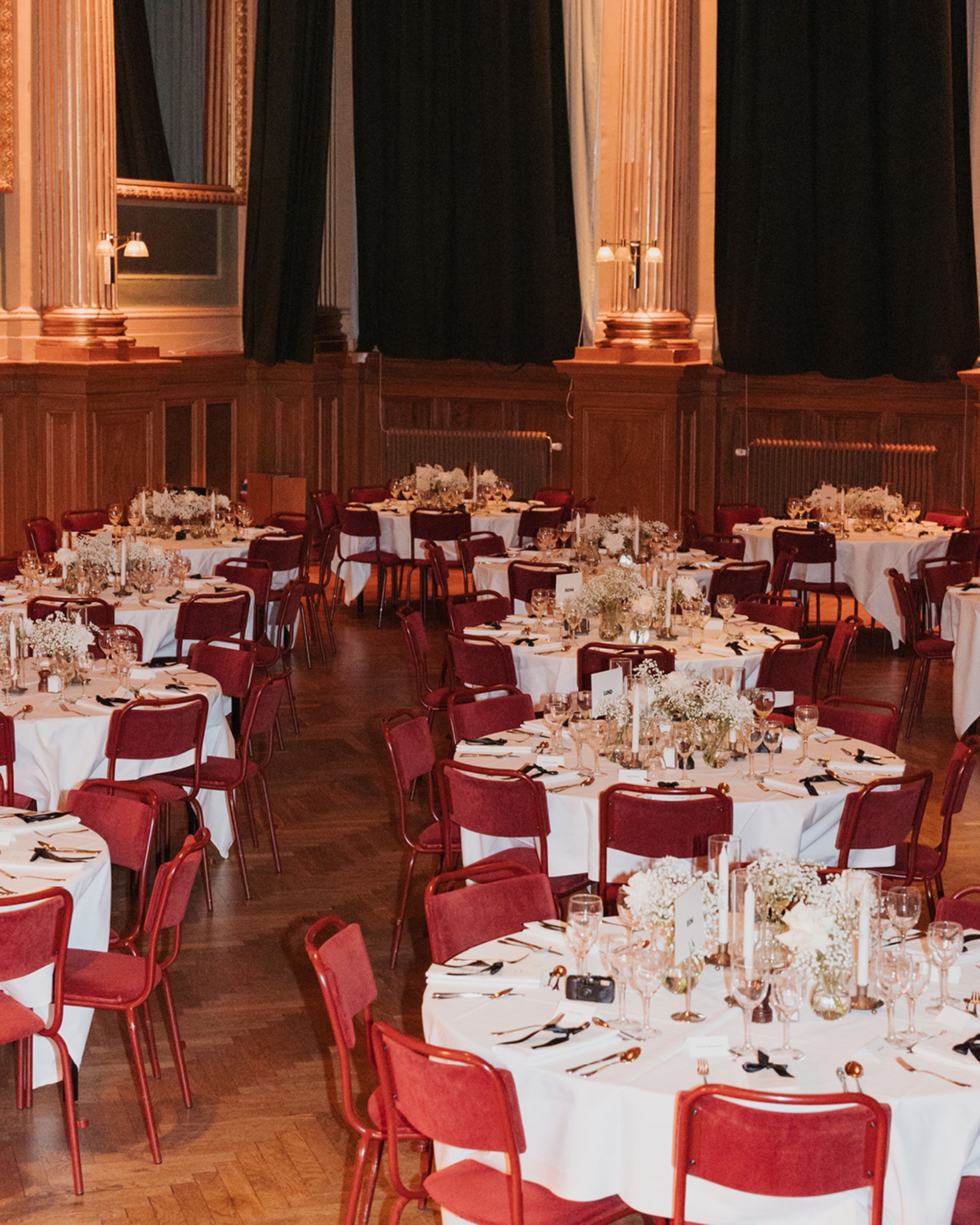 Elegant banquet hall with round tables set with white tablecloths, red chairs, glassware, and floral centerpieces under warm lighting.