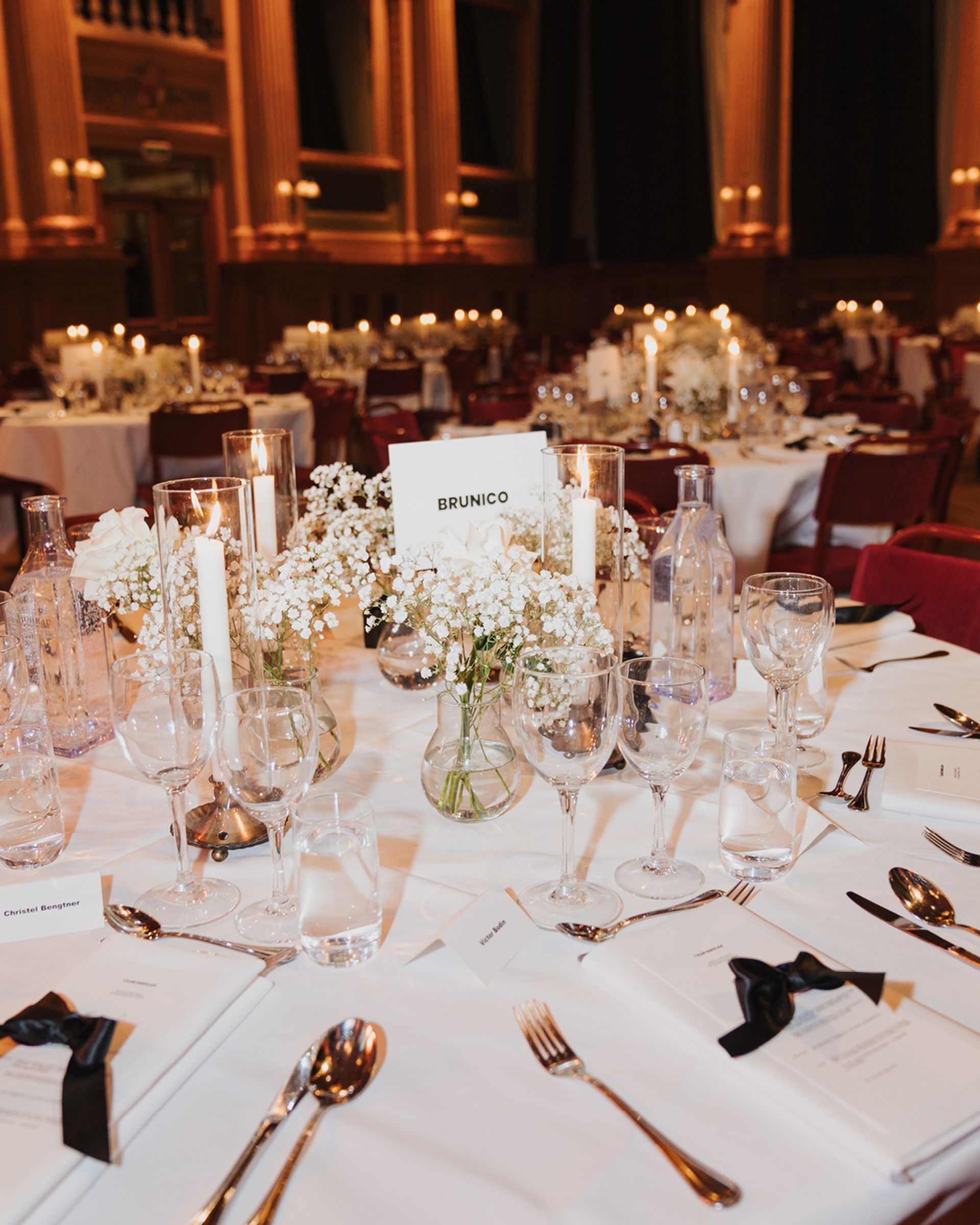 Elegant table setting with white flowers, candles, crystal glassware, and a "Brunico" card, in a grand banquet hall.