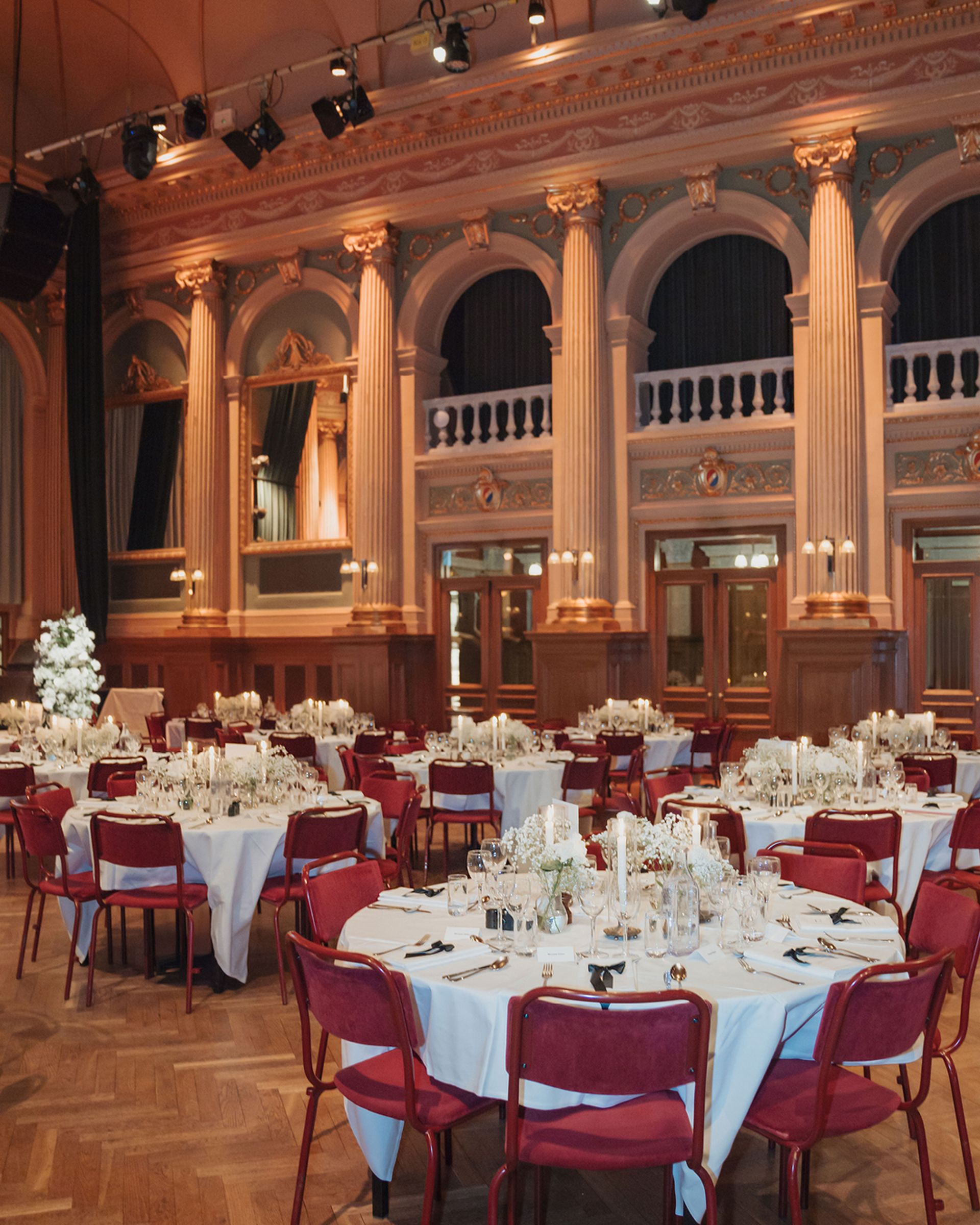 Elegant banquet hall with round tables set for an event, featuring red chairs, white tablecloths, and ornate columns under soft lighting.