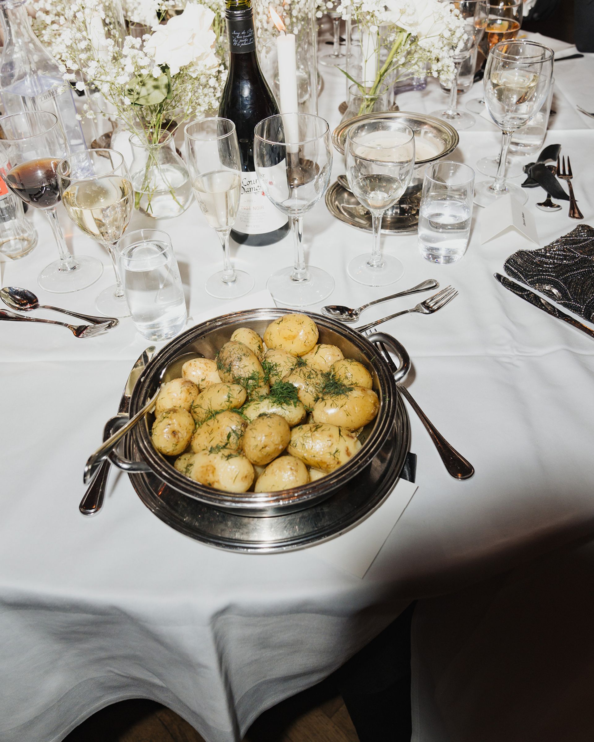 A bowl of boiled potatoes garnished with dill sits on a white tablecloth surrounded by wine glasses and floral centerpieces.