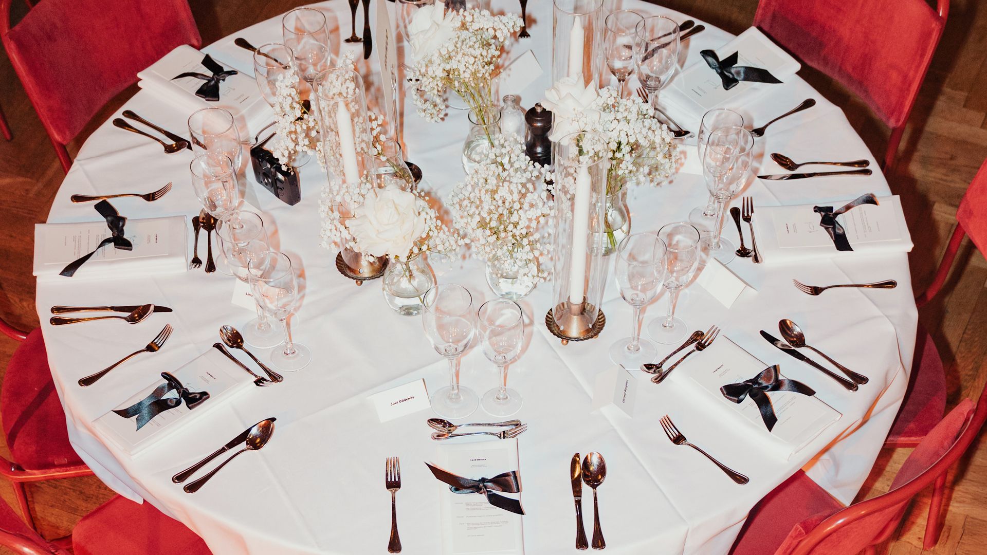 Elegant table setting with white tablecloth, red chairs, cutlery, glassware, black ribboned menus, and floral centerpieces featuring white roses and candles.