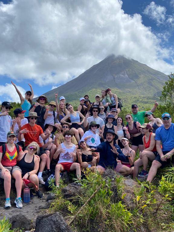Large group of students and leaders posing and cheering on a rocky outcrop with a cone-shaped volcano and clouds behind them in Costa Rica