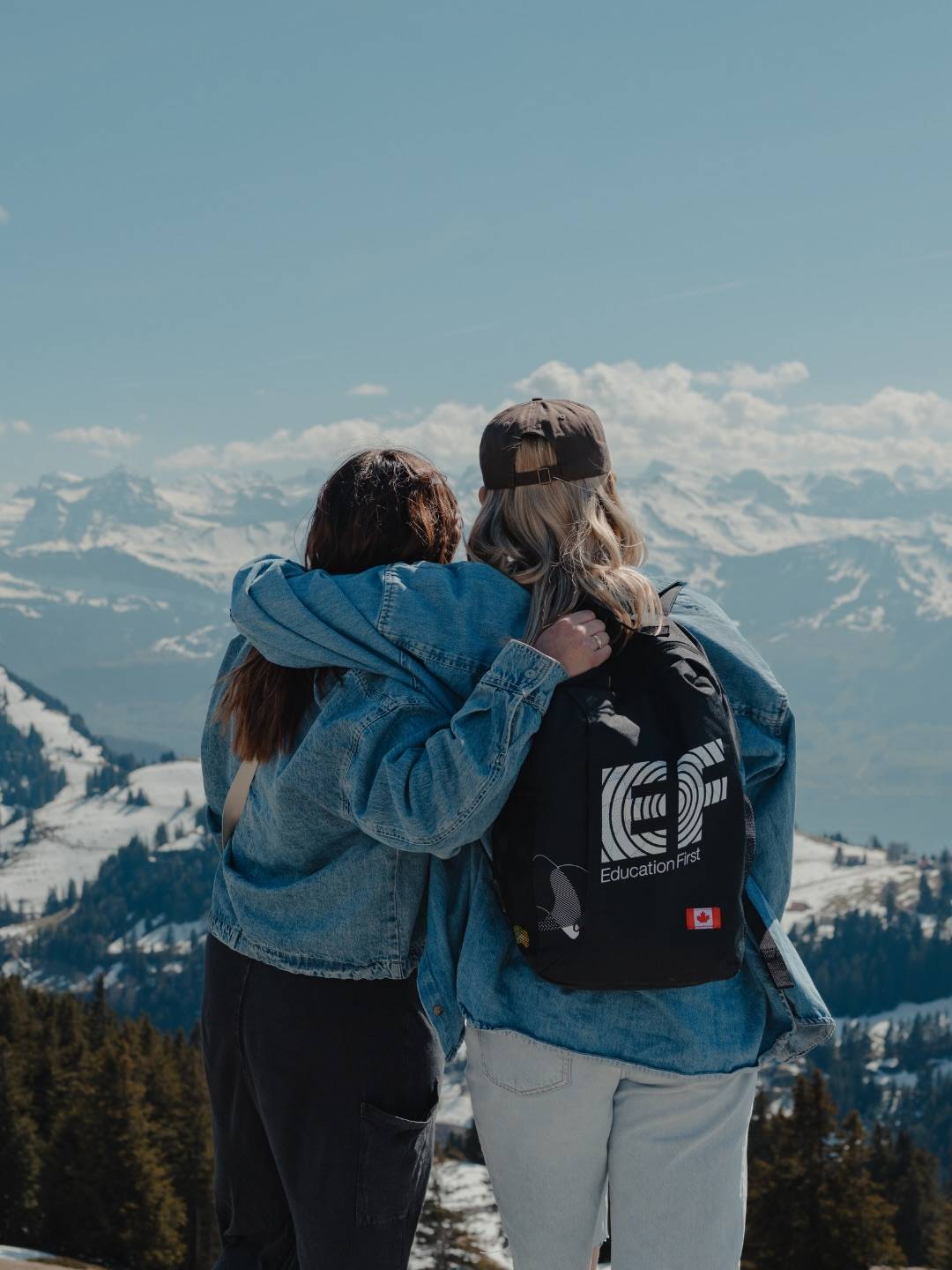 Two students seen from behind with their arms around each other, looking out at a snow-capped mountain range, one wearing an EF backpack with a Canadian flag patch