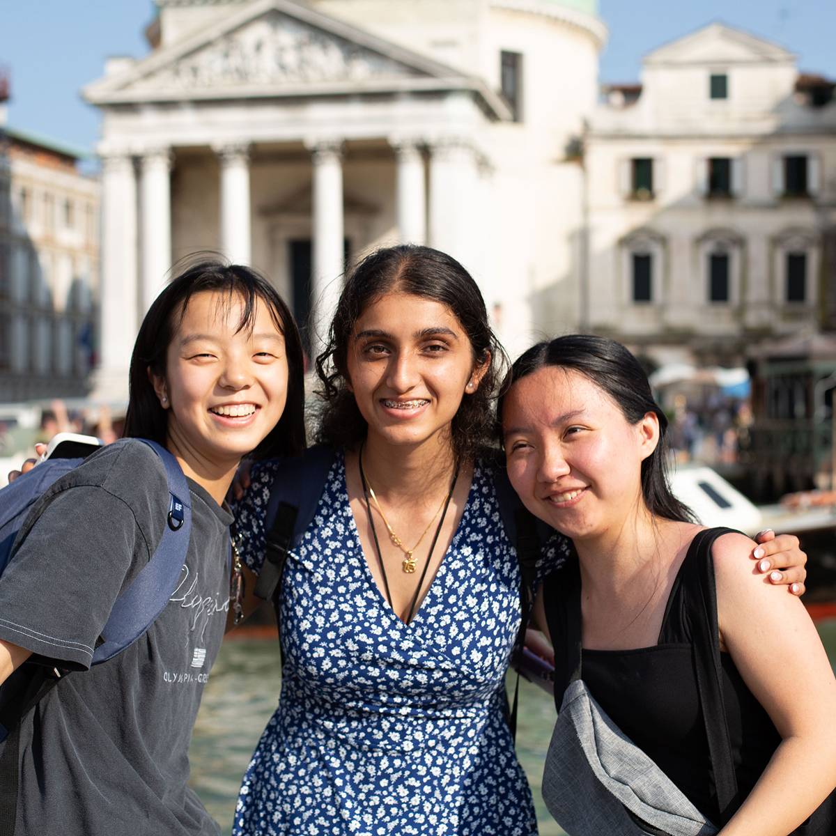 Three smiling students with their arms around each other beside the Grand Canal in Venice, with a neoclassical church with columns and a domed roof behind them