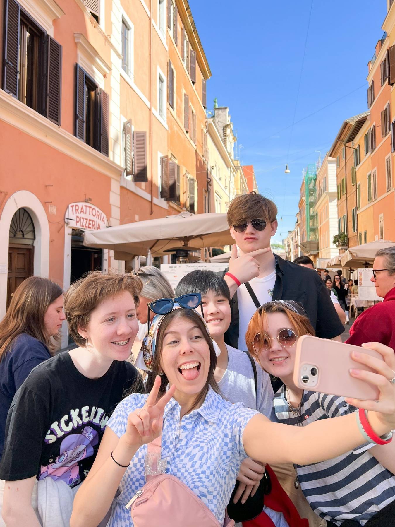Group of smiling students taking a selfie on a sunny street lined with orange and pink buildings, one making a peace sign and another wearing sunglasses on her head