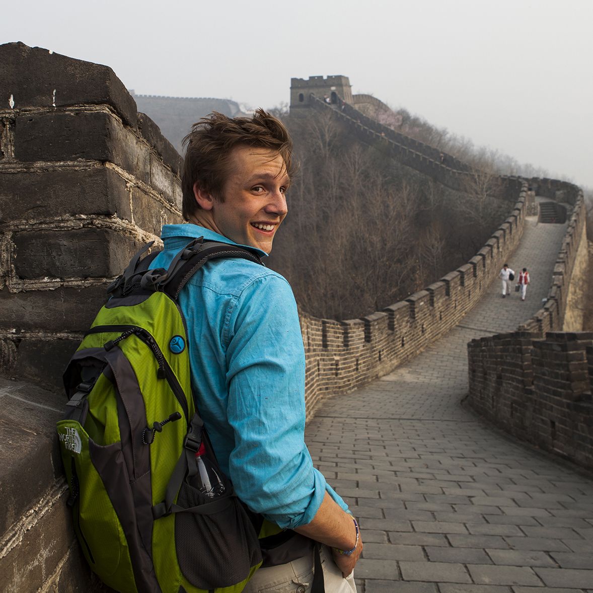 Smiling traveller in a teal jacket with a green backpack looking back over his shoulder while walking along a stone section of the Great Wall of China