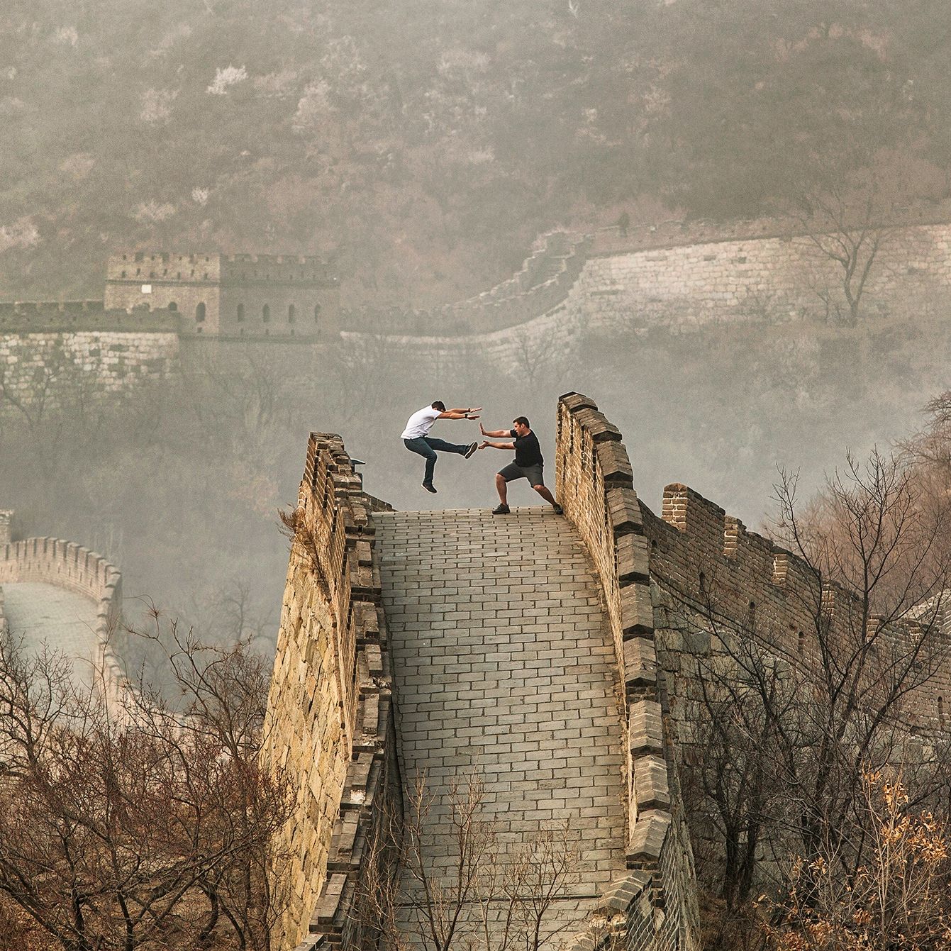 Two people striking a playful jumping pose on a stone walkway of the Great Wall of China, with watchtowers and misty hills in the background