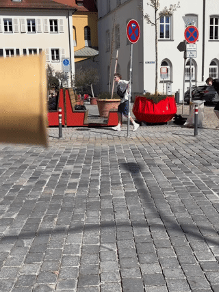 Video still of a paper cup held up at the left edge of the frame, with a few people walking across a cobblestone street in Europe behind it