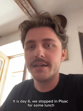 Clip of a man with tousled hair and a moustache in a black T-shirt speaking to the camera indoors, with the caption "It is day 6, we stopped in Pisac for some lunch"