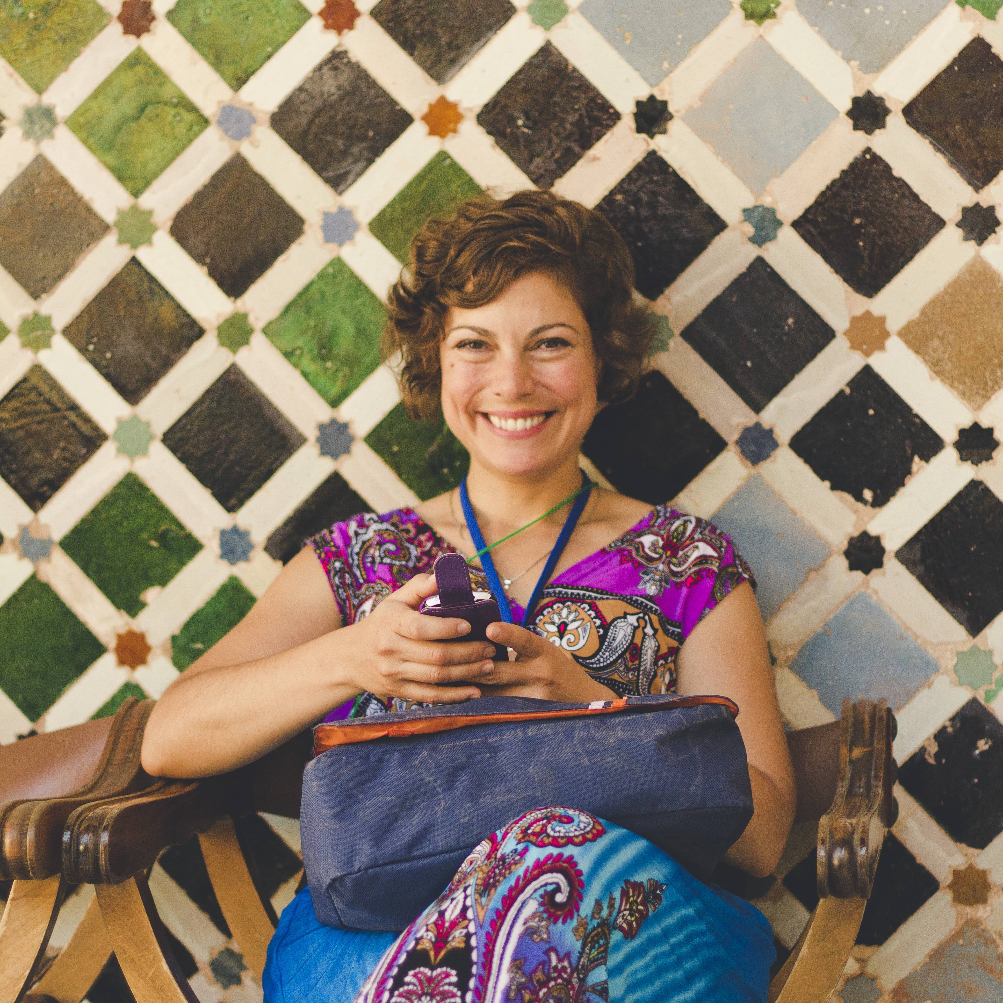 Smiling woman in a colourful patterned top seated on a wooden chair holding a phone in front of a wall of green, brown and cream geometric tiles in Spain