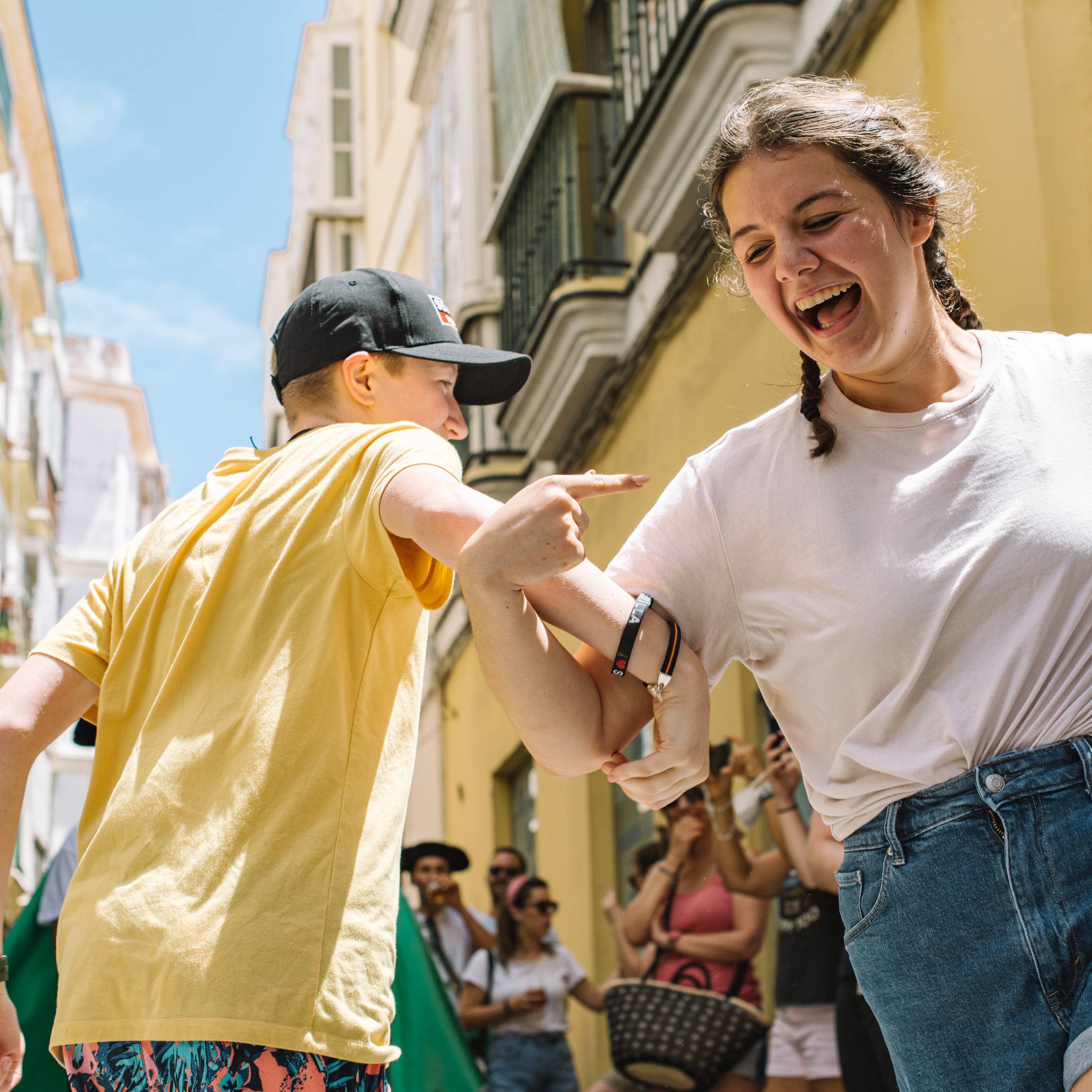 Two people laughing and pointing at each other, one in a yellow shirt and cap and one with a braid and white T-shirt, on a sunny narrow street lined with yellow buildings in Spain