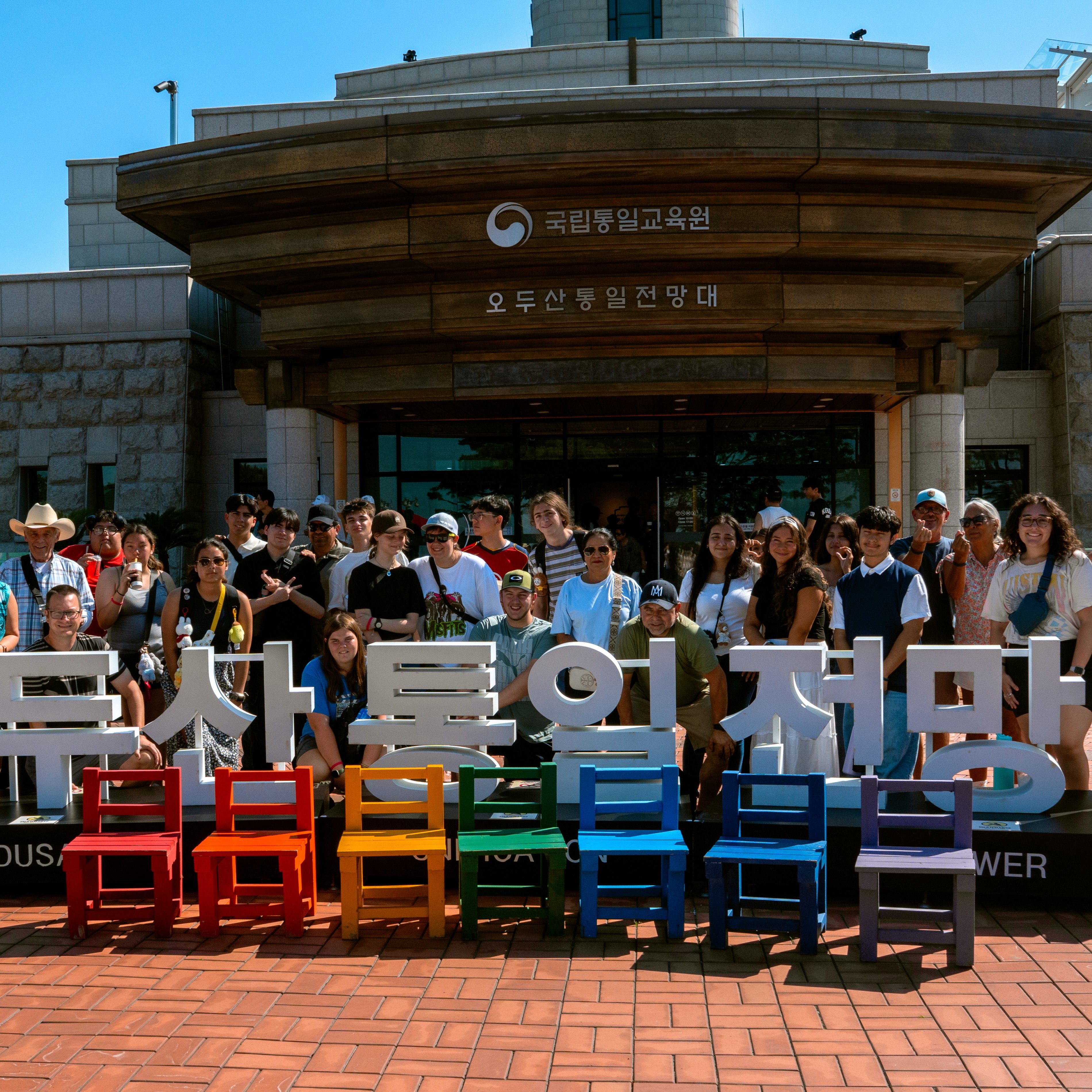 Large group of students and teachers posing on colourful chairs spelling out Korean lettering in front of an observatory building in South Korea