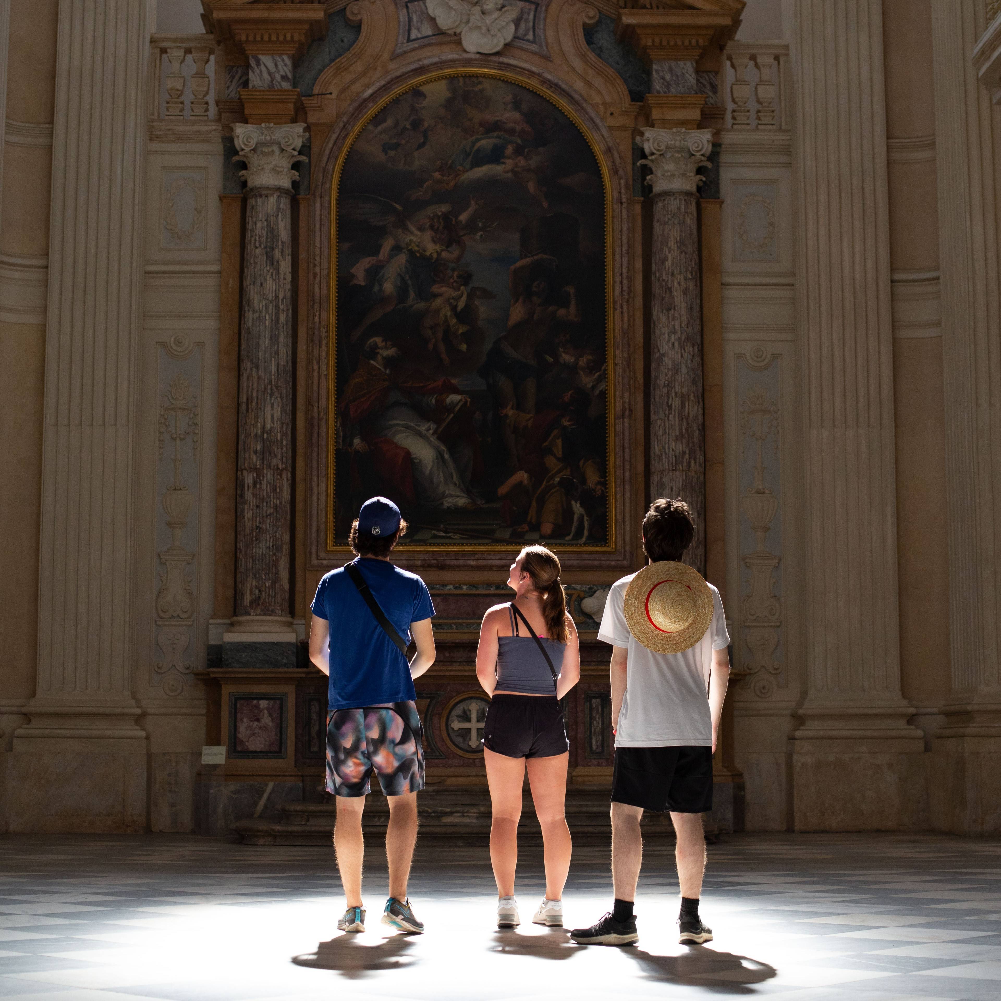 Three students seen from behind standing in a sunlit hall, looking up at a large painting in a gilded arch framed by marble columns in Italy