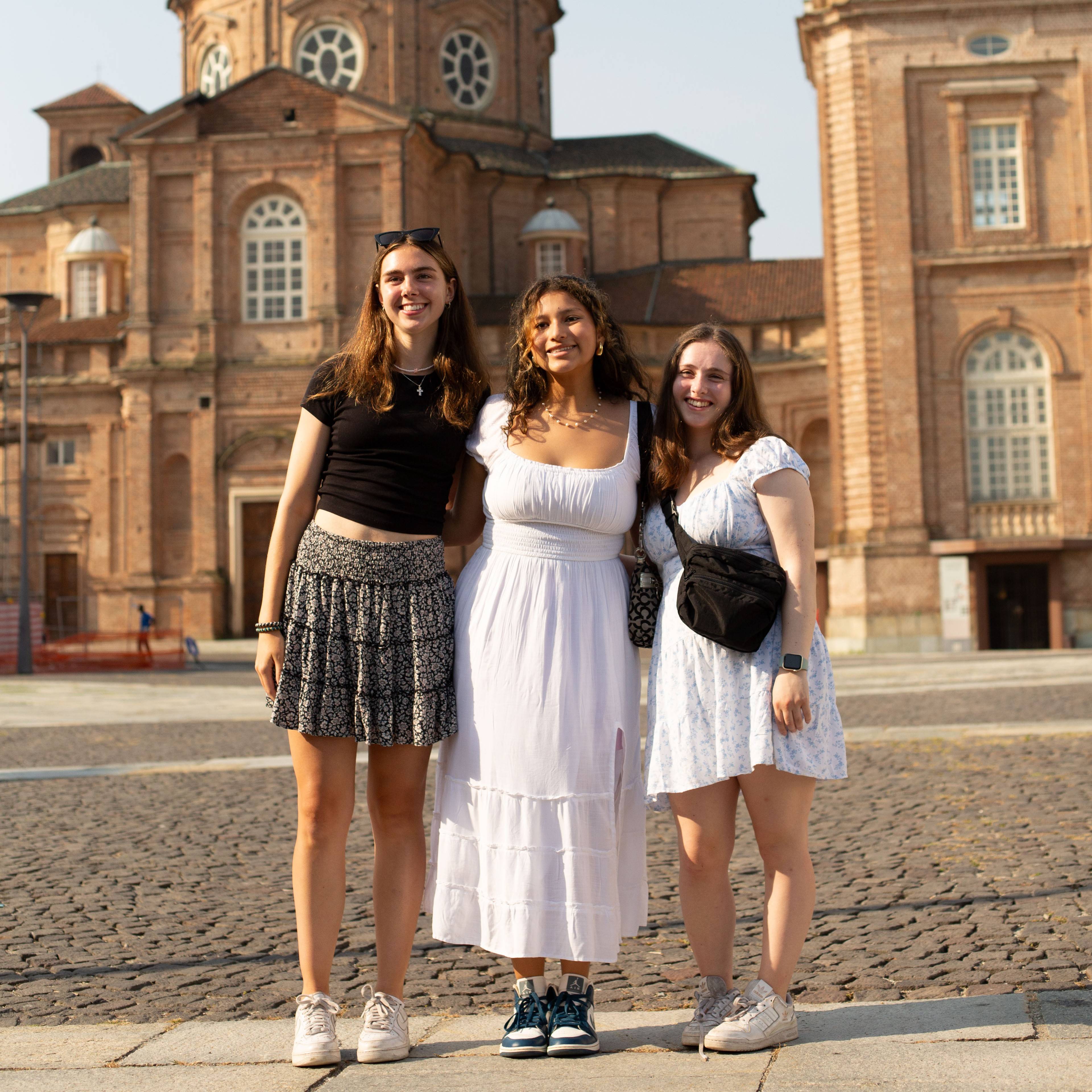 Three smiling students standing arm in arm on a cobblestone square in front of a large Baroque brick palace with a clock tower in Italy