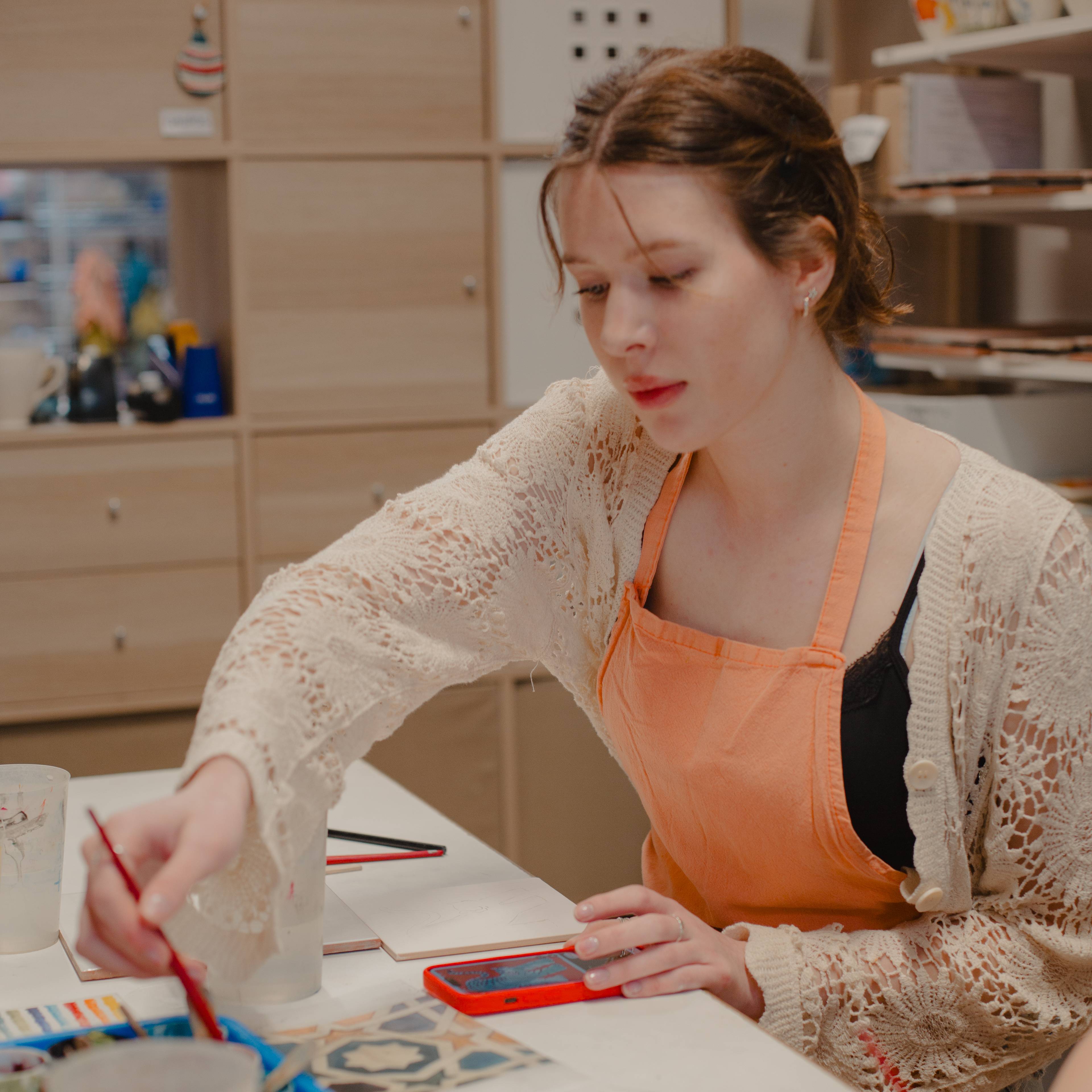 Woman in an orange apron painting with a red brush at a workshop table with paints and craft supplies, with wooden shelves behind her in Spain