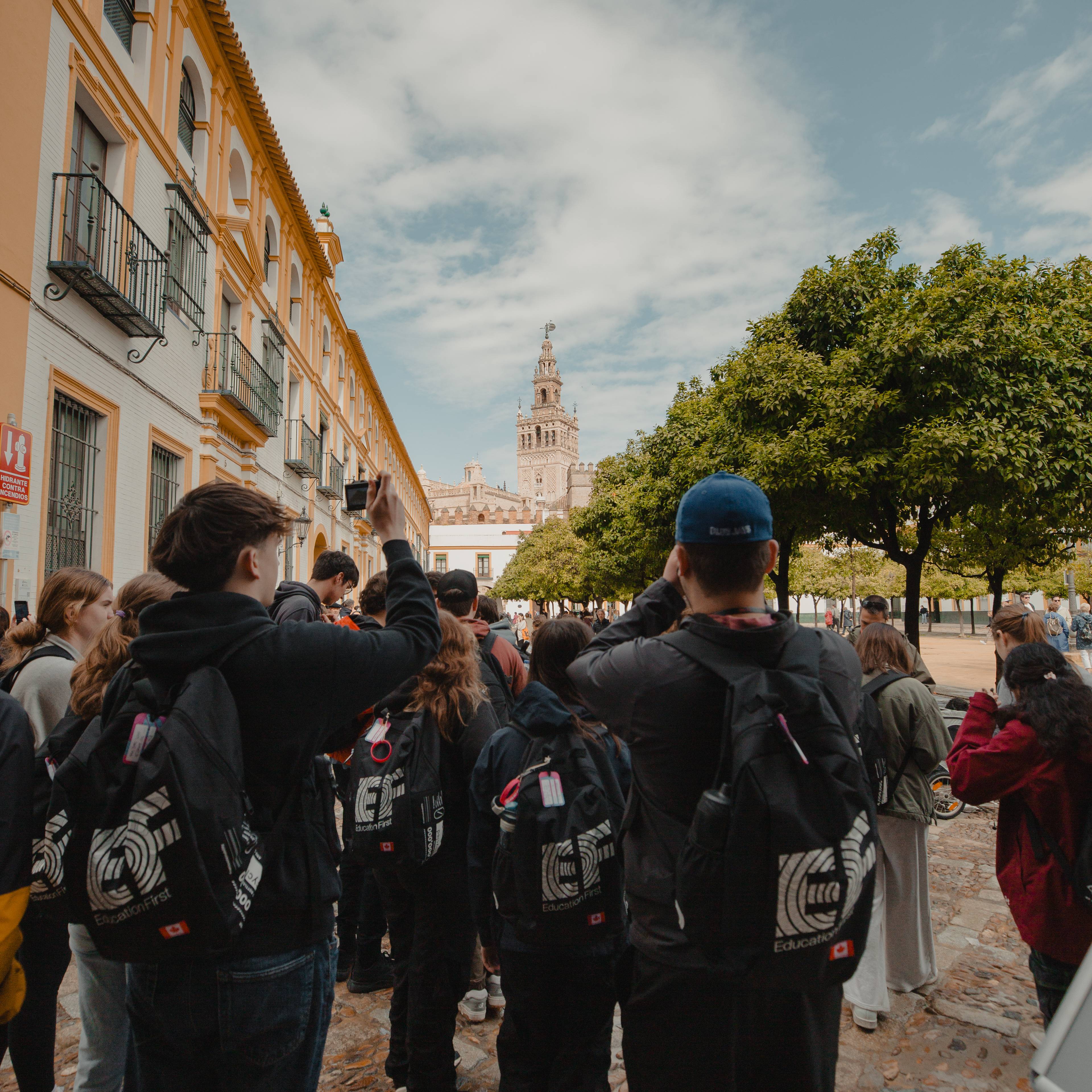 Group of students wearing EF backpacks seen from behind on a historic street lined with orange trees and a church tower in the distance in Seville, Spain