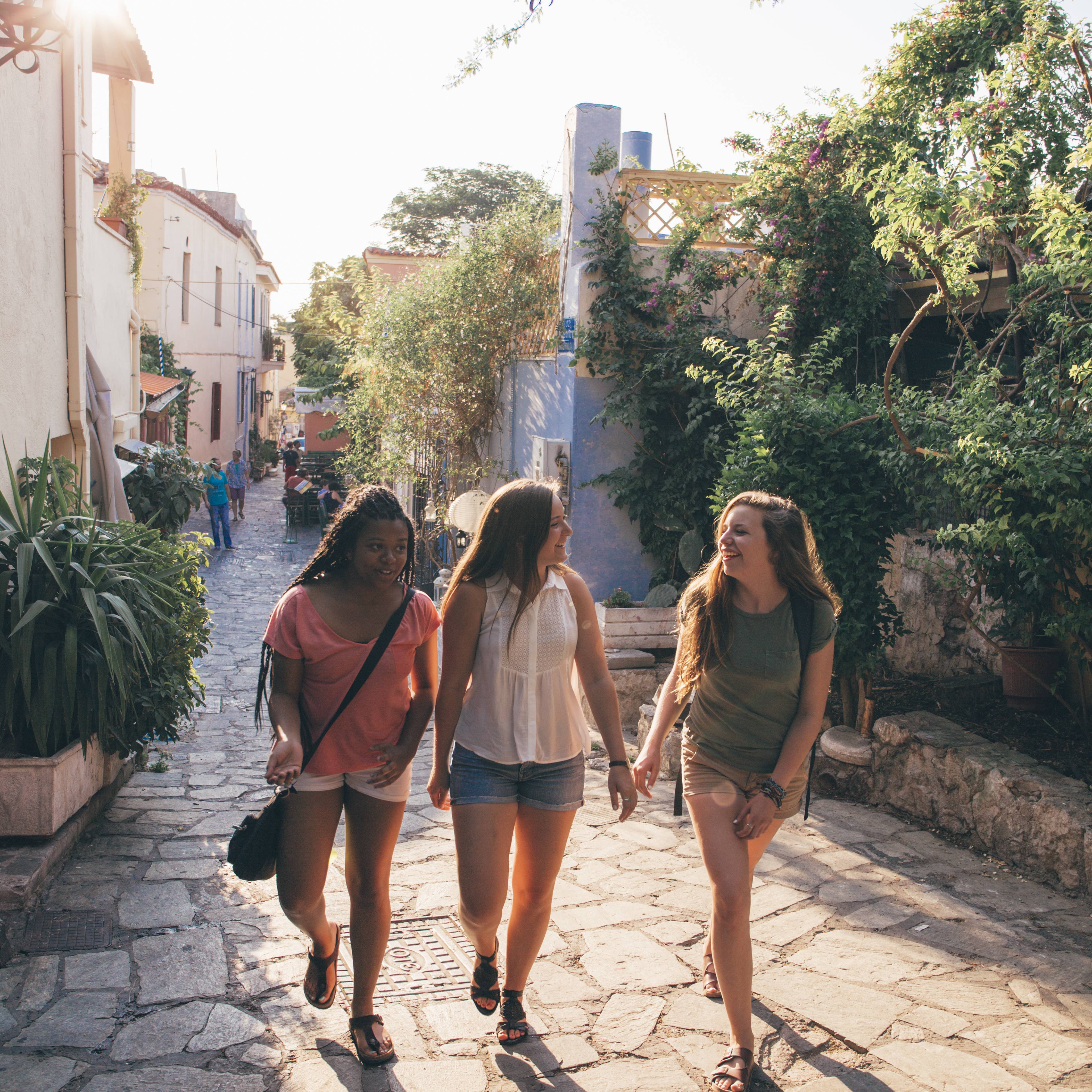 Three students walking together down a sunny cobblestone lane lined with whitewashed buildings, green plants and trees in Greece