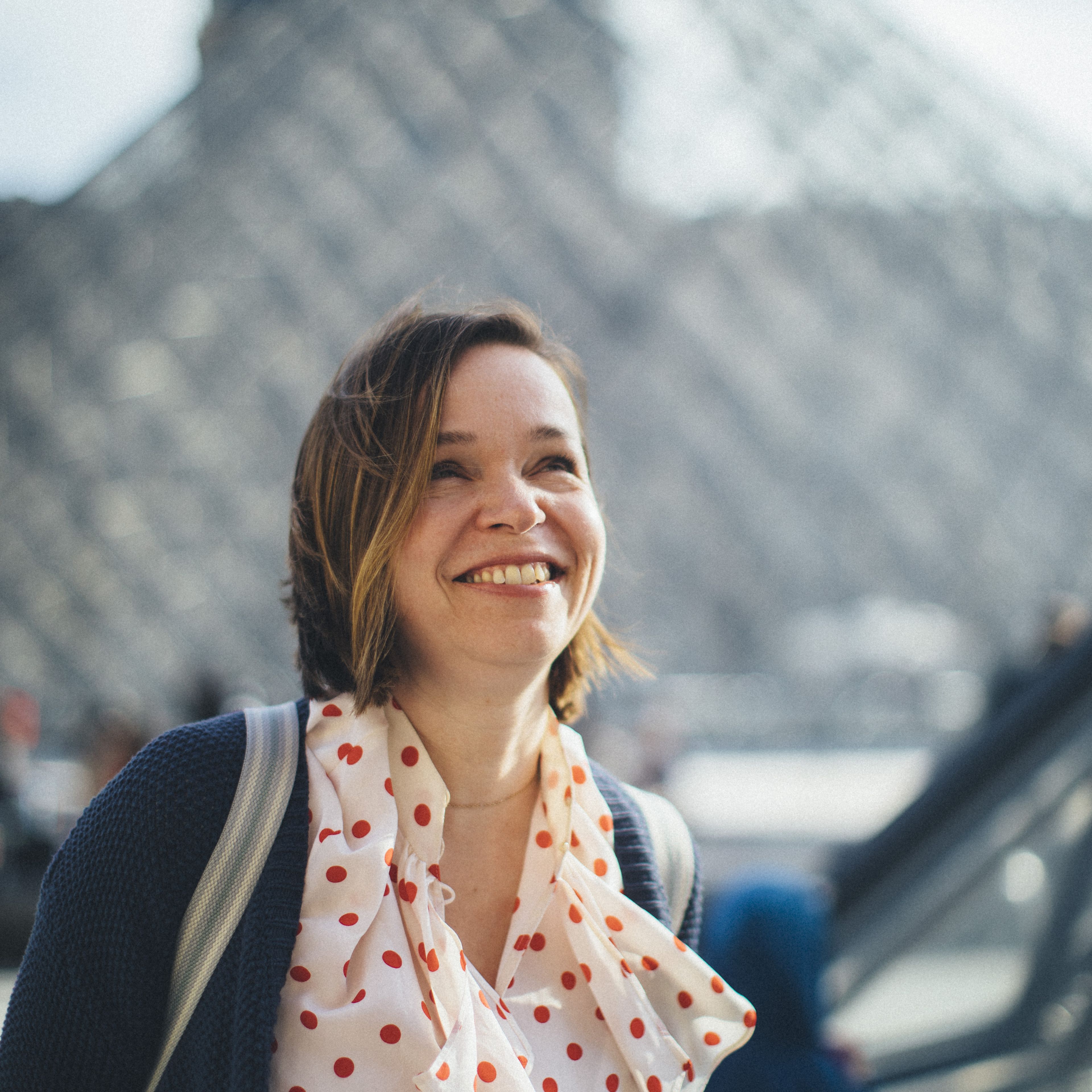 Smiling woman with a white-and-red polka dot scarf and a backpack strap, with the glass pyramid of the Louvre blurred behind her in Paris, France