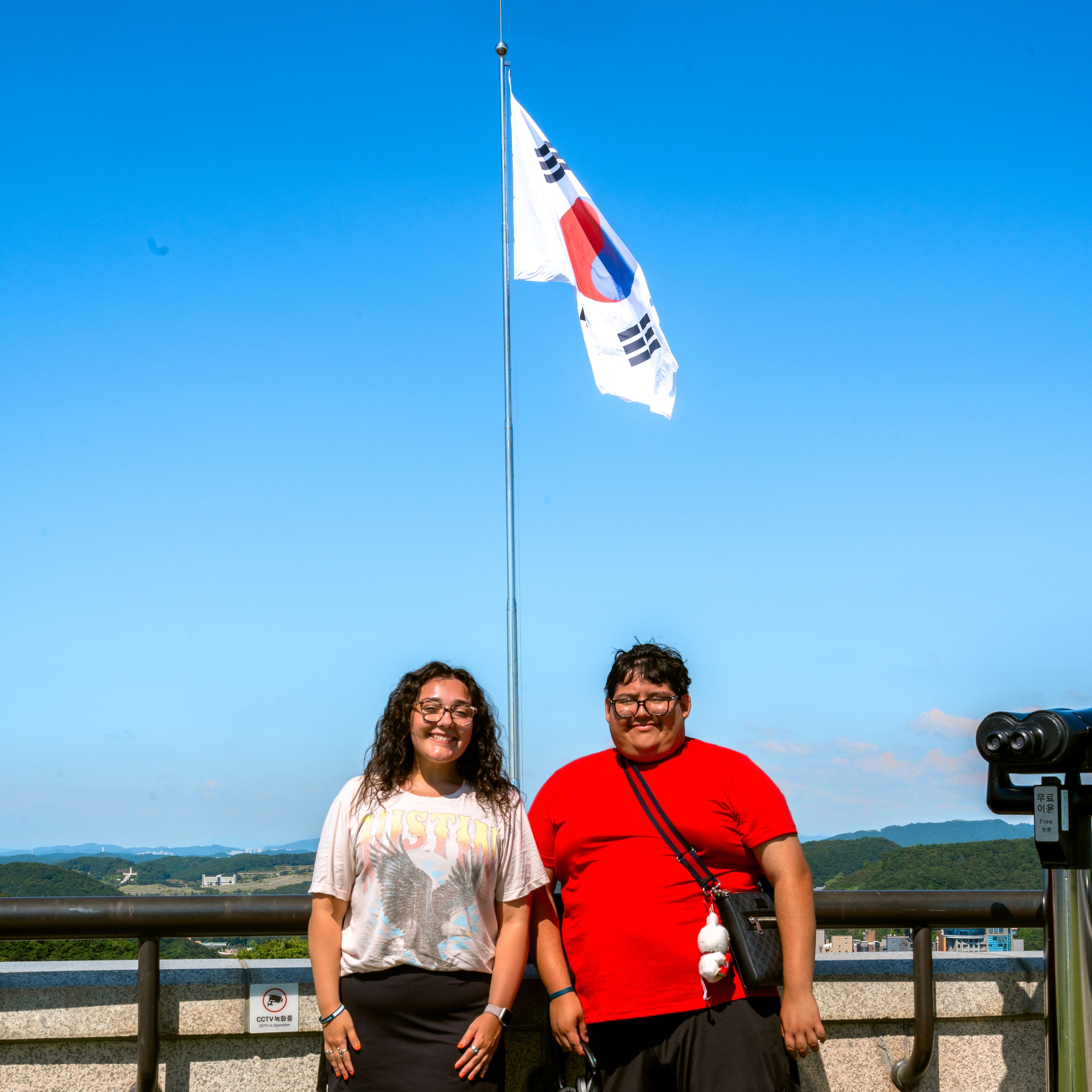 Two smiling people standing at a lookout beneath the South Korean flag flying against a clear blue sky, with hills in the distance