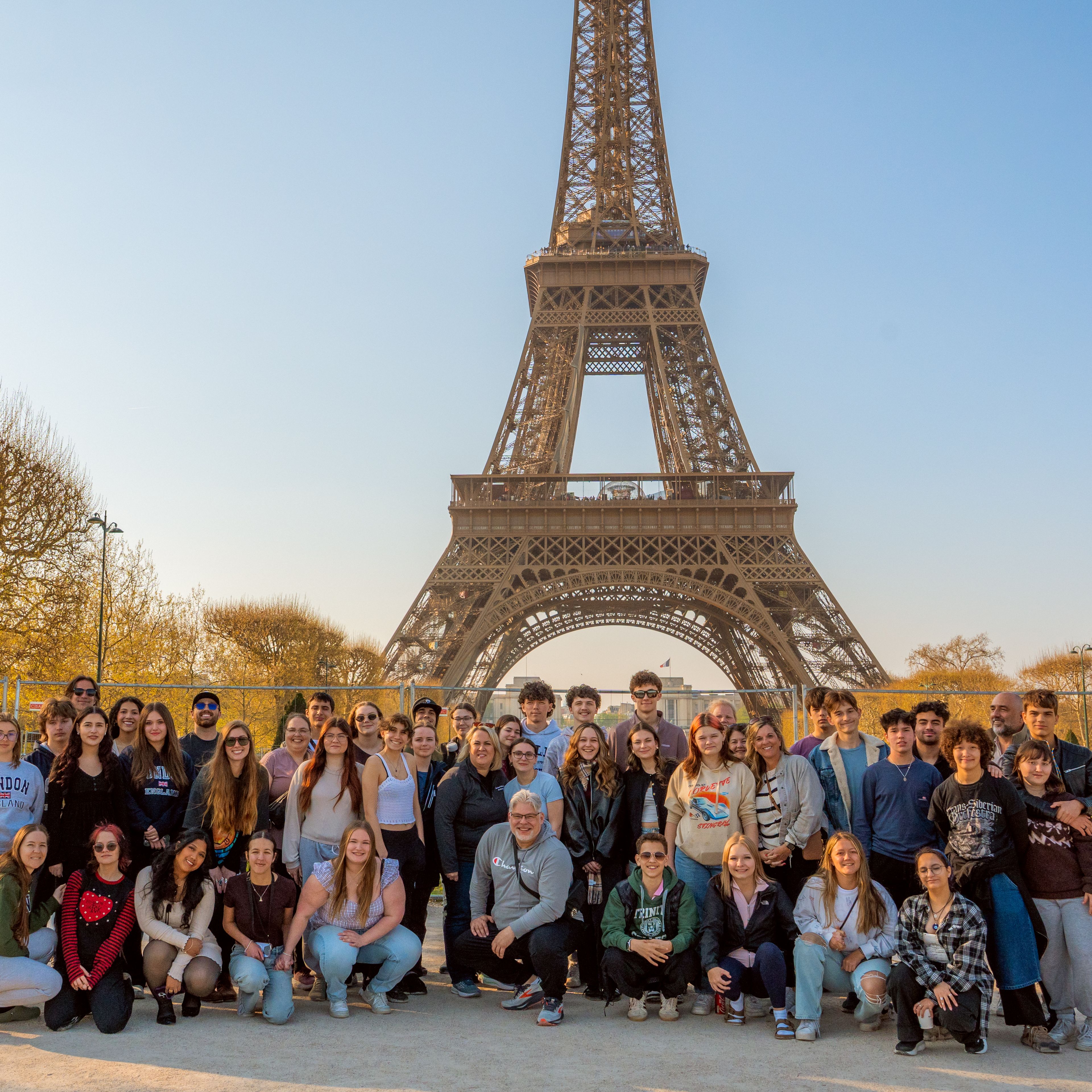 Large group of students and teachers posing together on the ground in front of the Eiffel Tower in Paris, France, on a sunny day