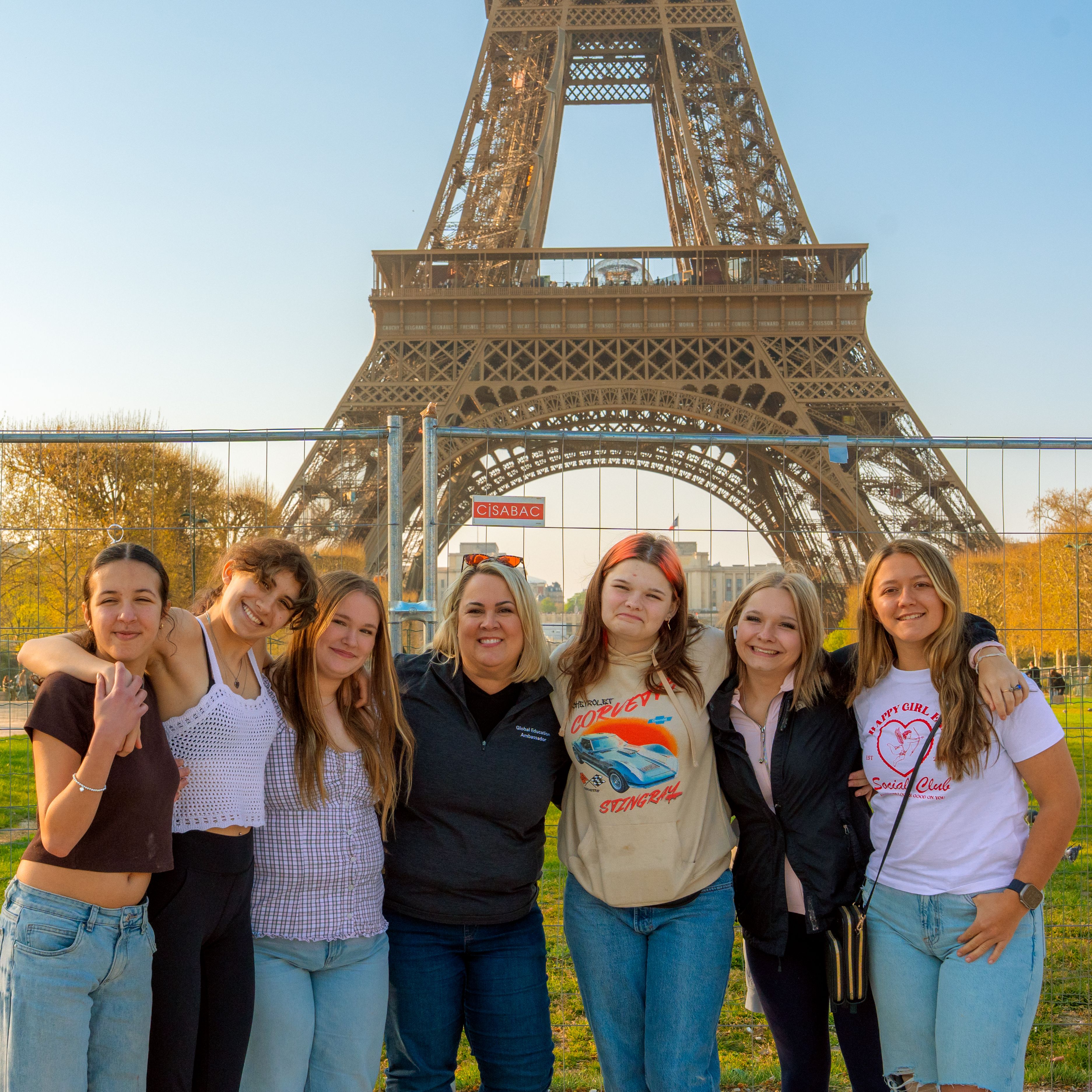 Six smiling students and their teacher standing arm in arm on the grass in front of the Eiffel Tower in Paris, France, on a sunny day