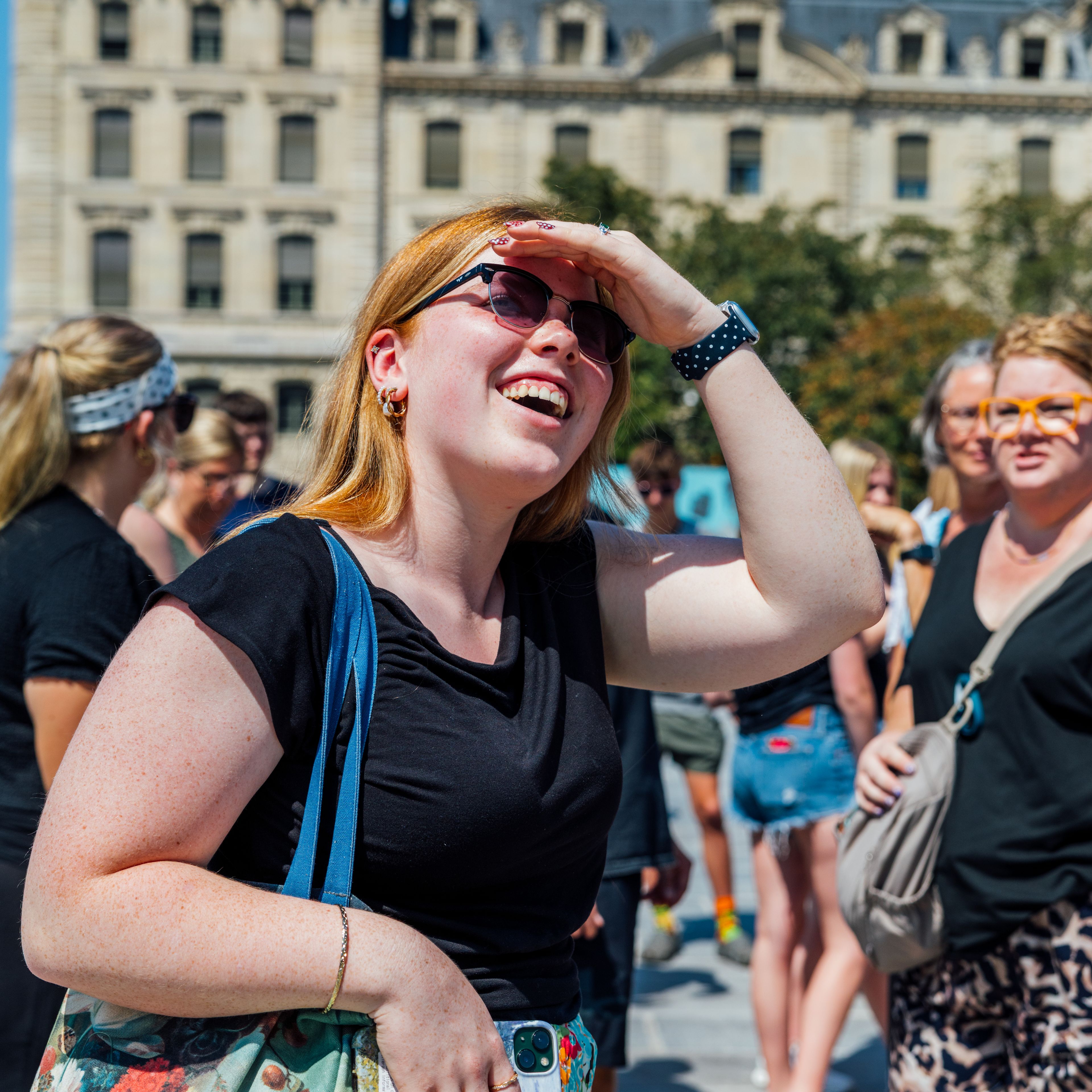 Laughing woman in sunglasses shading her eyes from the sun, with other travellers beside her and classic cream-coloured Paris buildings behind them