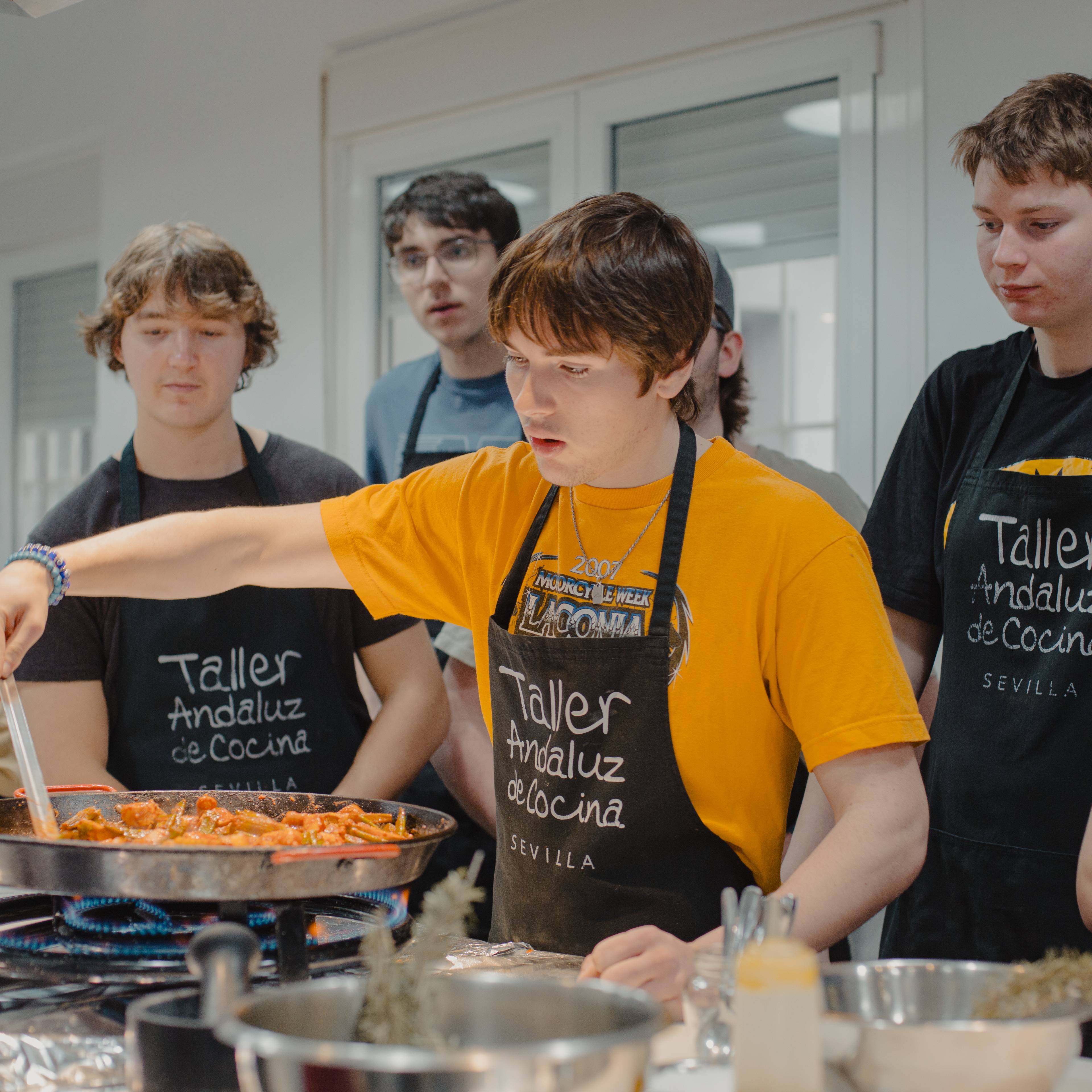 Students in aprons reading "Taller Andaluz" gathered around a large pan of paella, one in a yellow shirt stirring, during a cooking class in Spain