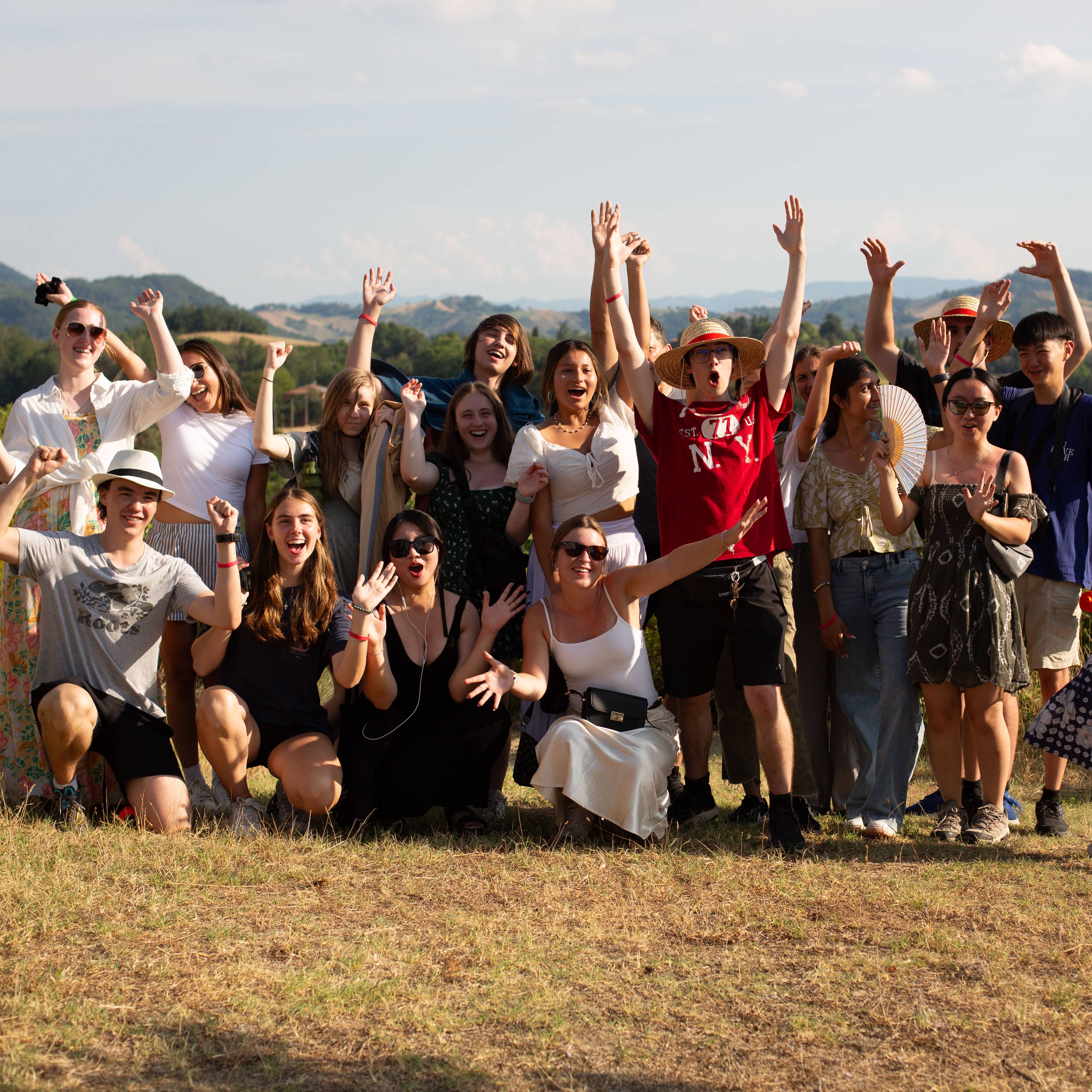 Large group of students cheering with their arms raised on a grassy hilltop at golden hour, with rolling hills in the background in Italy