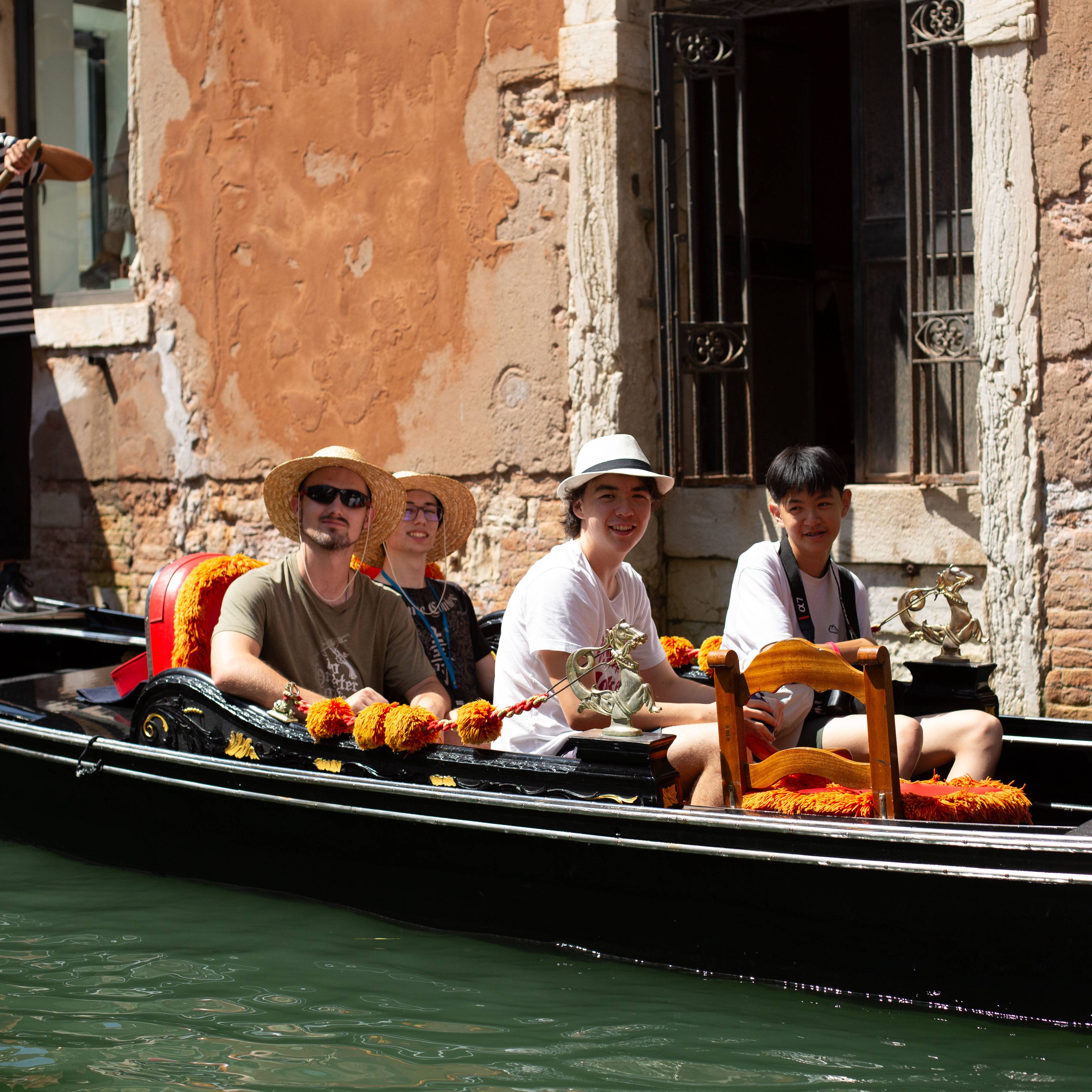 Four travellers in sun hats and sunglasses sitting in a black gondola with orange cushions on a green canal beside a weathered Venetian building in Italy