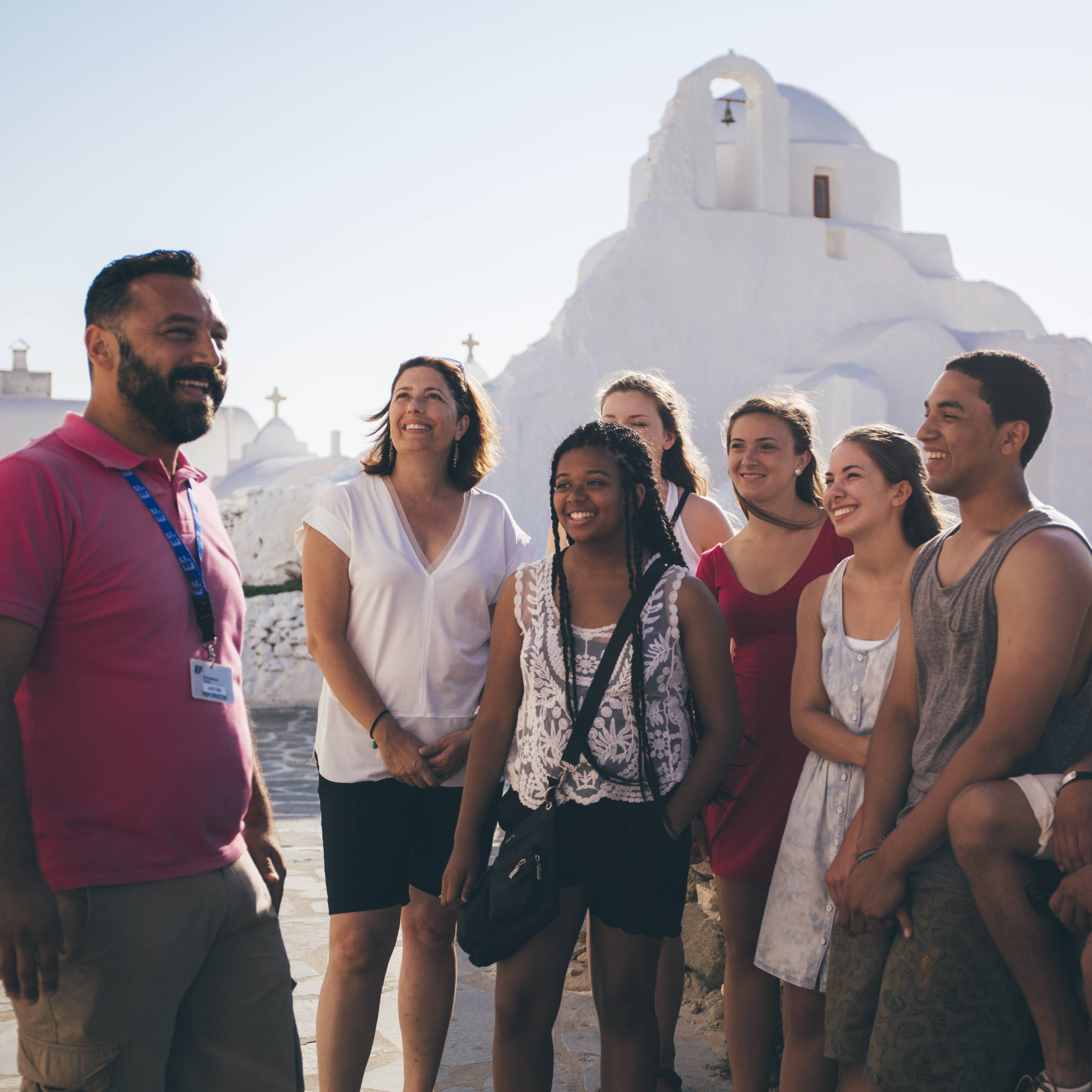 Man in a pink polo shirt and lanyard smiling at a group of students seated and standing in front of a whitewashed Greek church with a bell tower