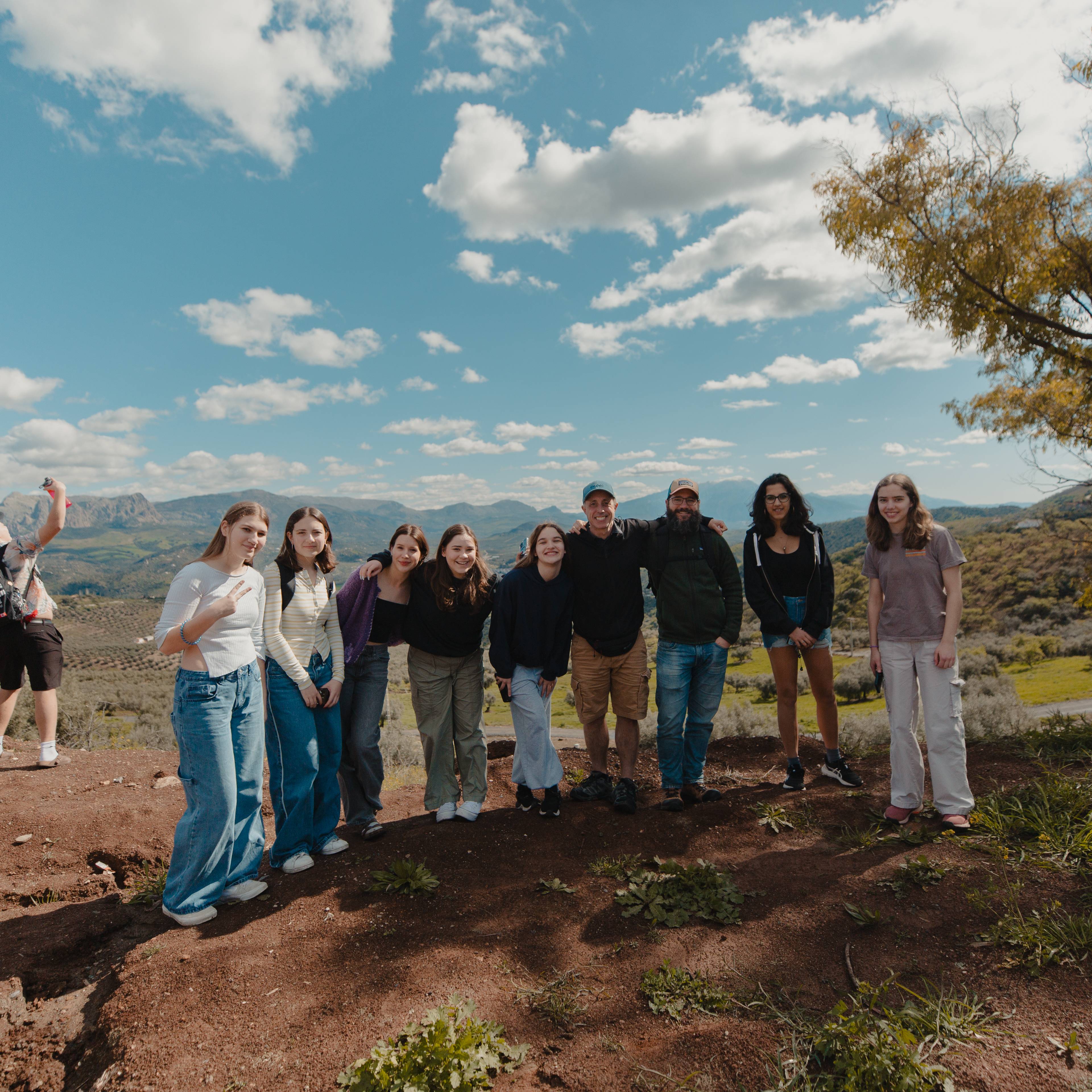 Group of students standing in a line on a dry hillside with a view over a valley and mountains under a partly cloudy sky in Spain