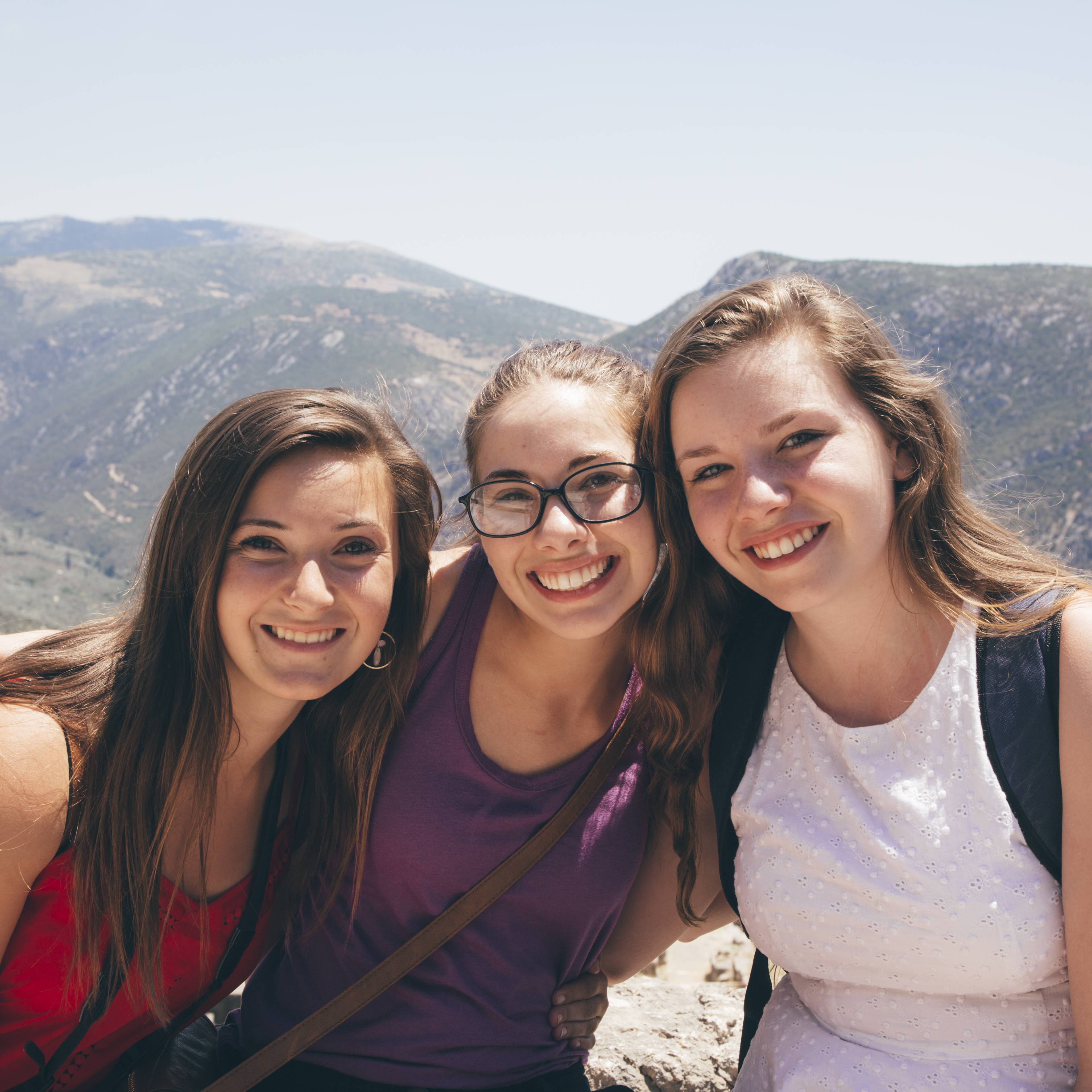 Three smiling students leaning together for a photo with a sunlit mountain landscape behind them in Greece
