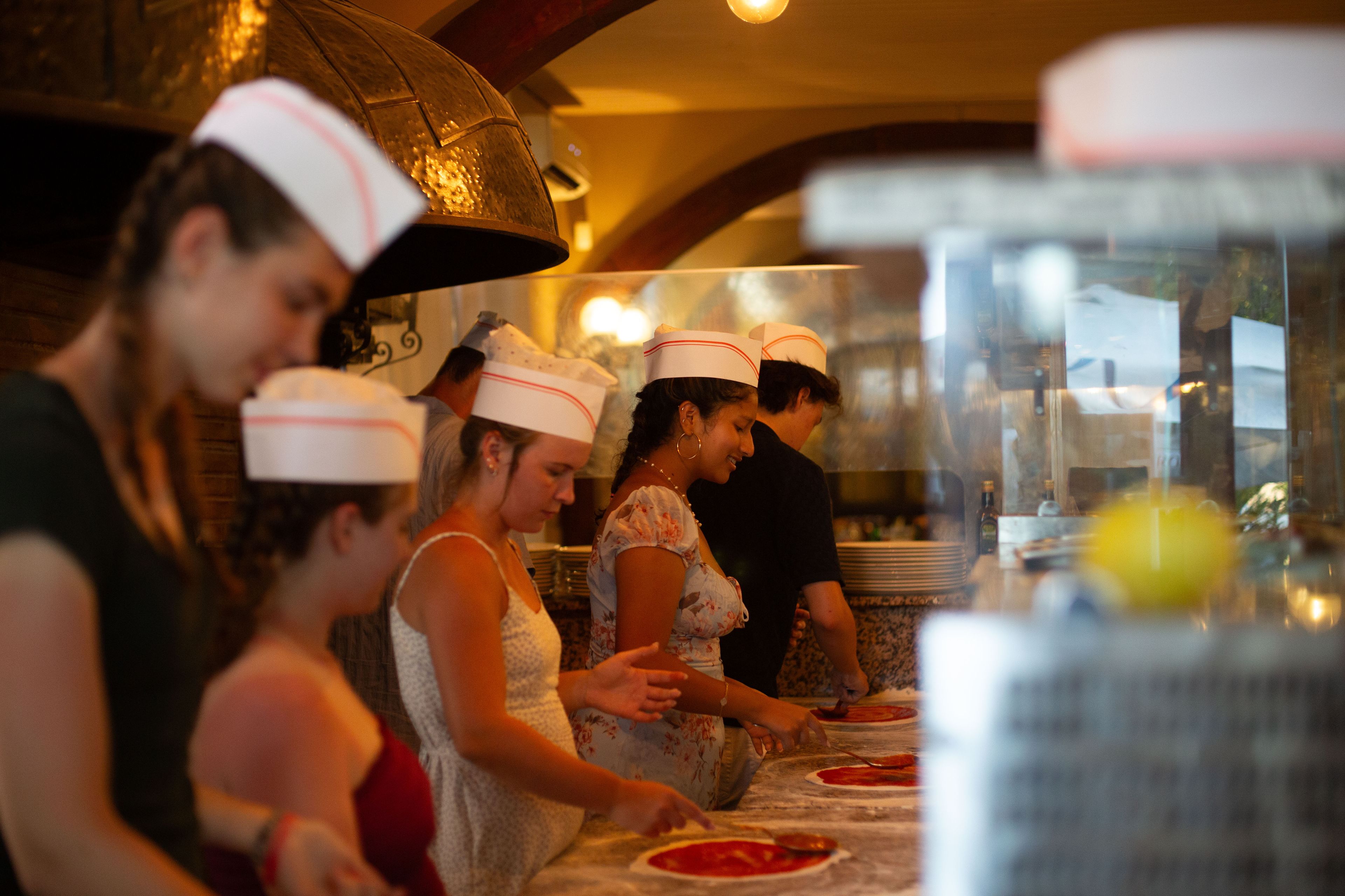 Students wearing white paper chef hats gathered around a kitchen counter during a pizza-making class in Italy