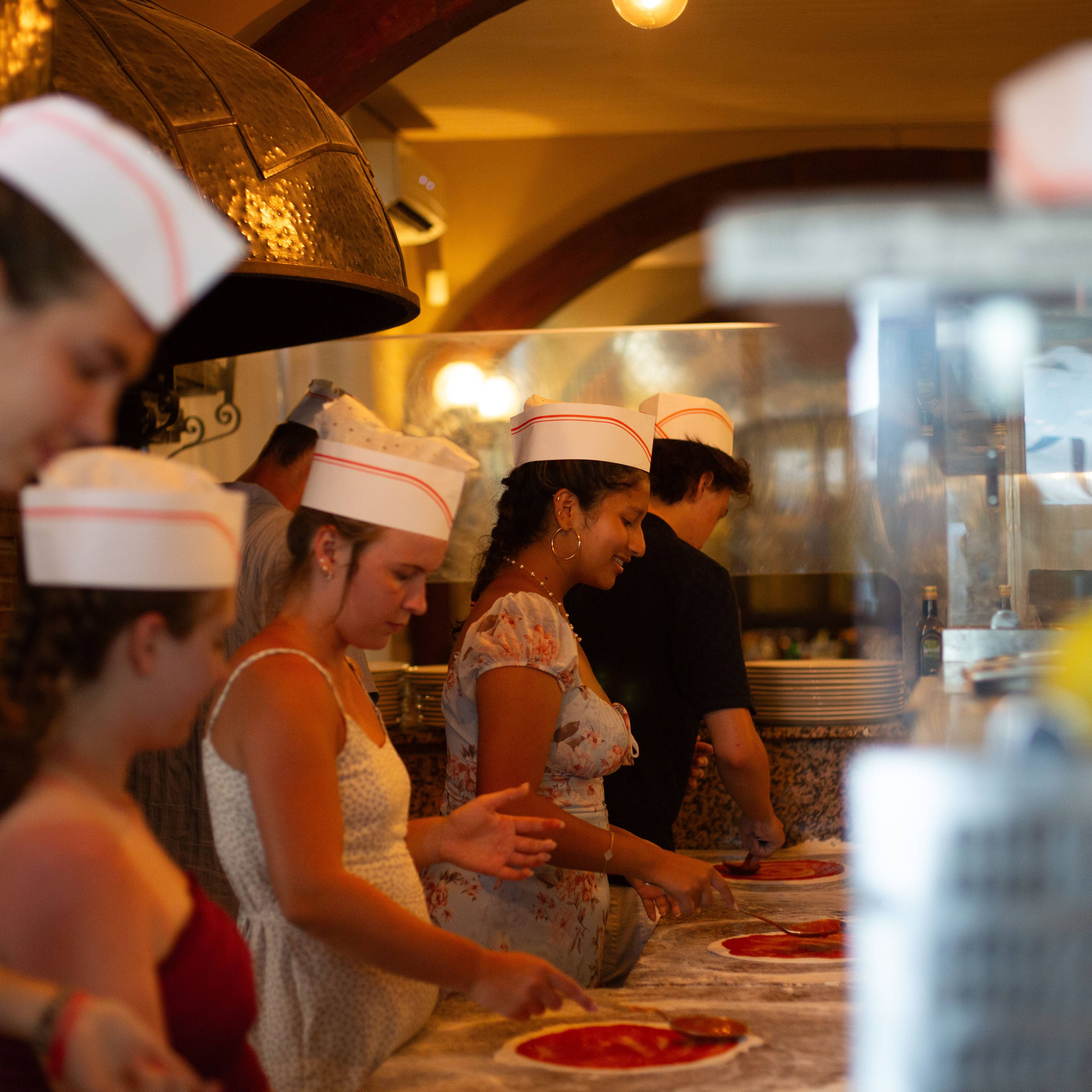 Students wearing white paper chef hats gathered around a kitchen counter during a pizza-making class in Italy
