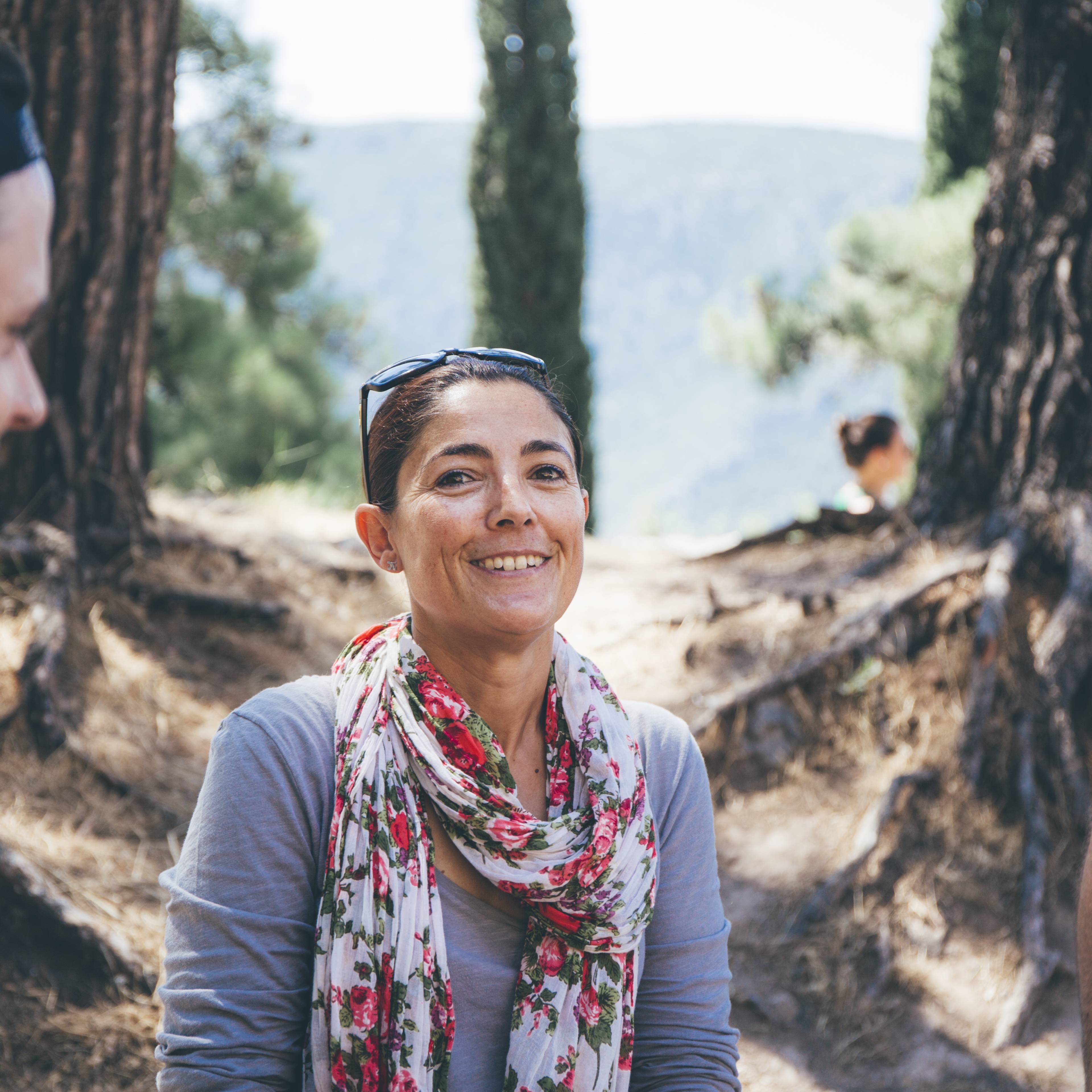 Smiling woman with sunglasses on her head and a floral scarf standing on a path among tall cypress trees, with hazy hills in the distance in Greece