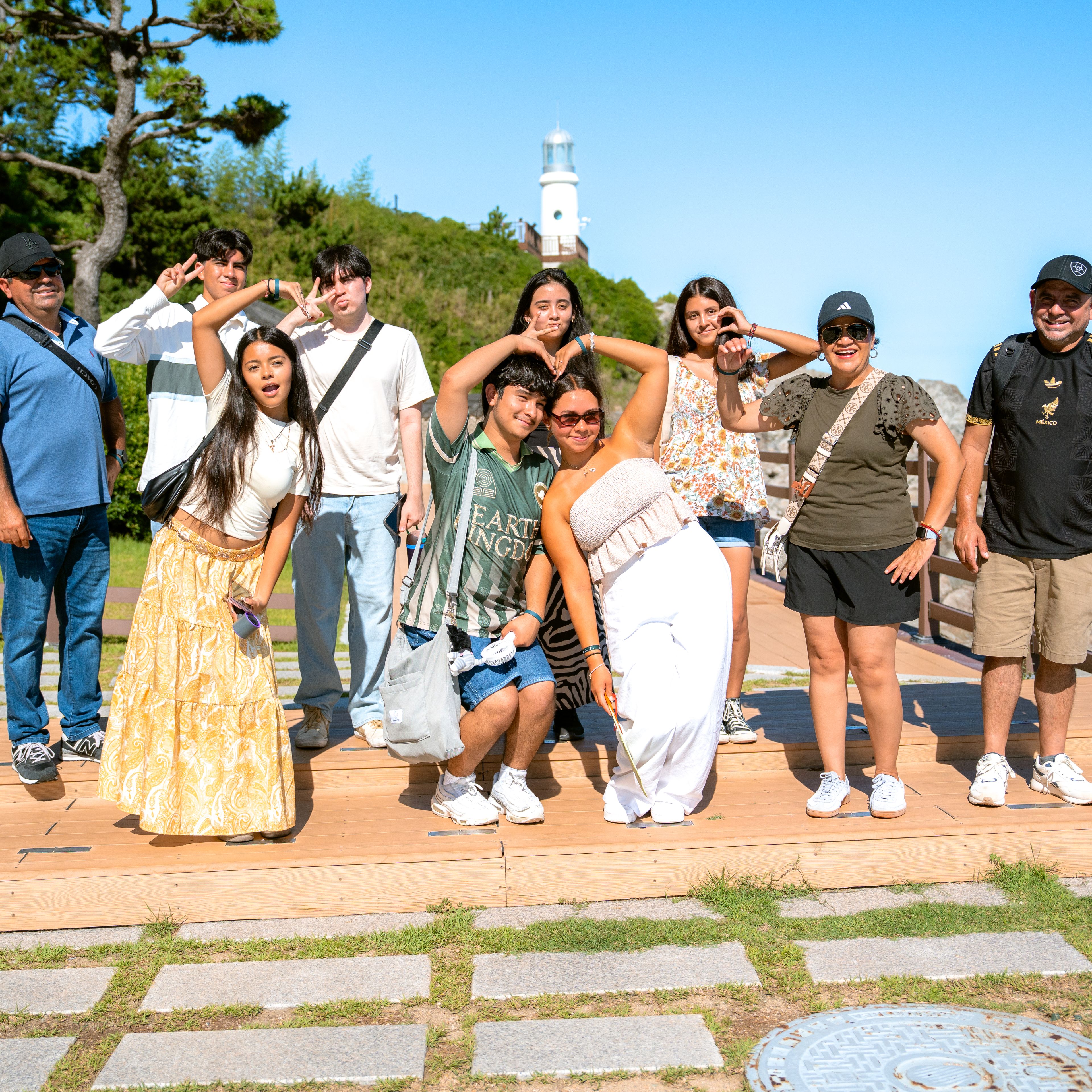 Group of students striking playful poses on a wooden viewing deck, with a white lighthouse on a green hillside behind them in South Korea