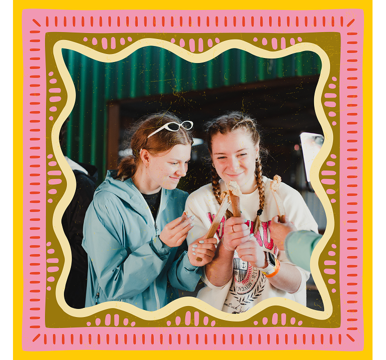 Two smiling students holding pieces of sugarcane, one in a blue rain jacket and one with braids, inside a decorative patterned frame in Costa Rica