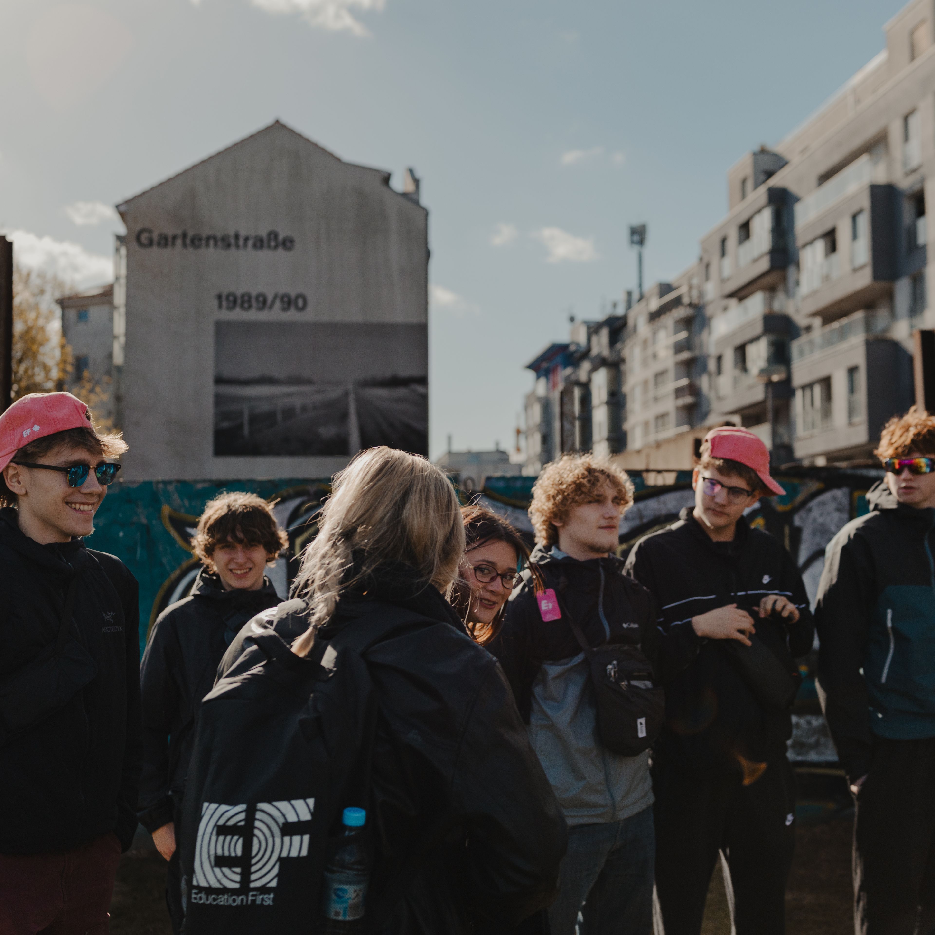 Group of students listening to a guide in front of a graffiti-covered wall and a building mural marked "Gartenstraße 1989/90" in Berlin, Germany