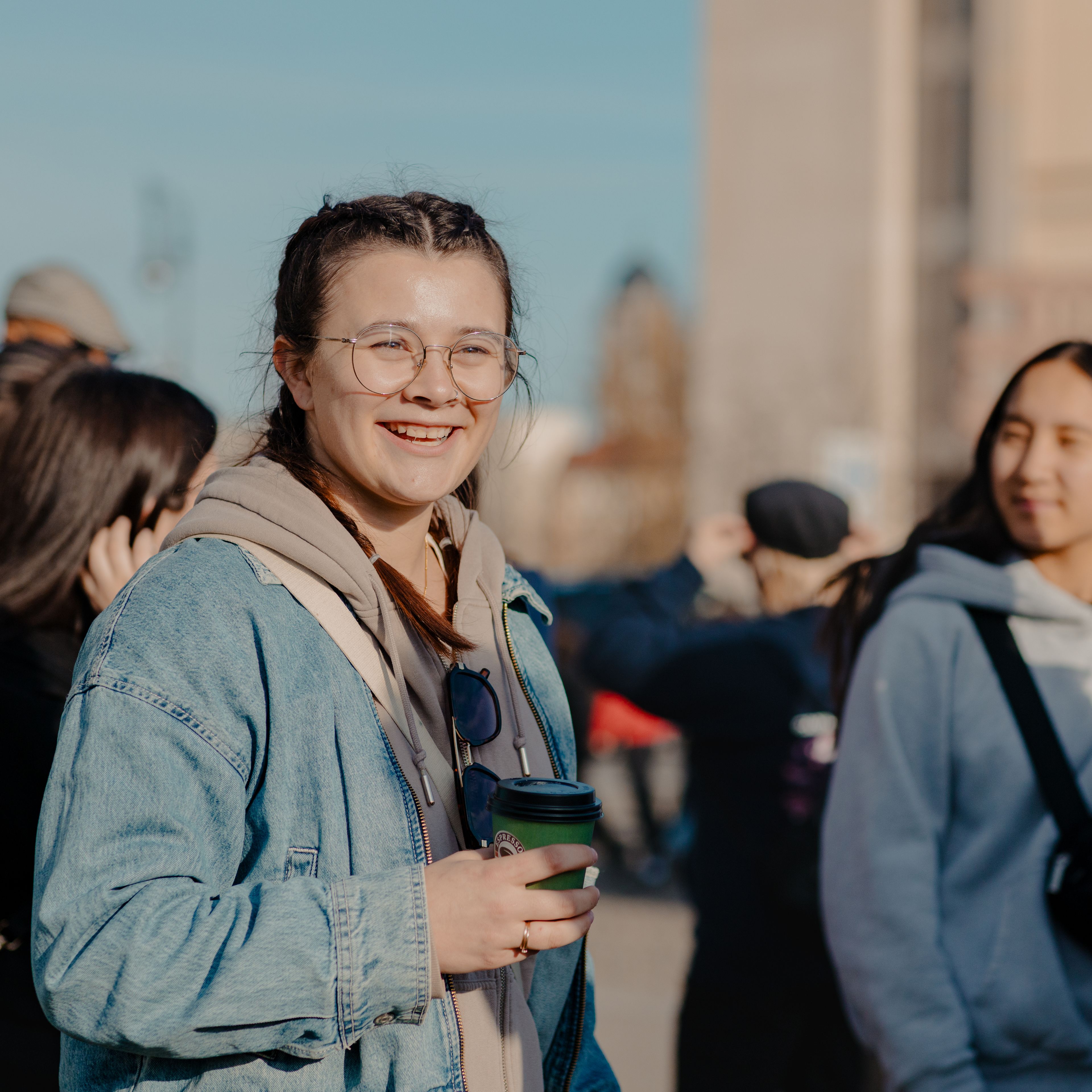 Smiling teacher with braids and glasses, wearing a denim jacket and holding a coffee cup, with students gathered behind her on an outdoor group tour in Germany